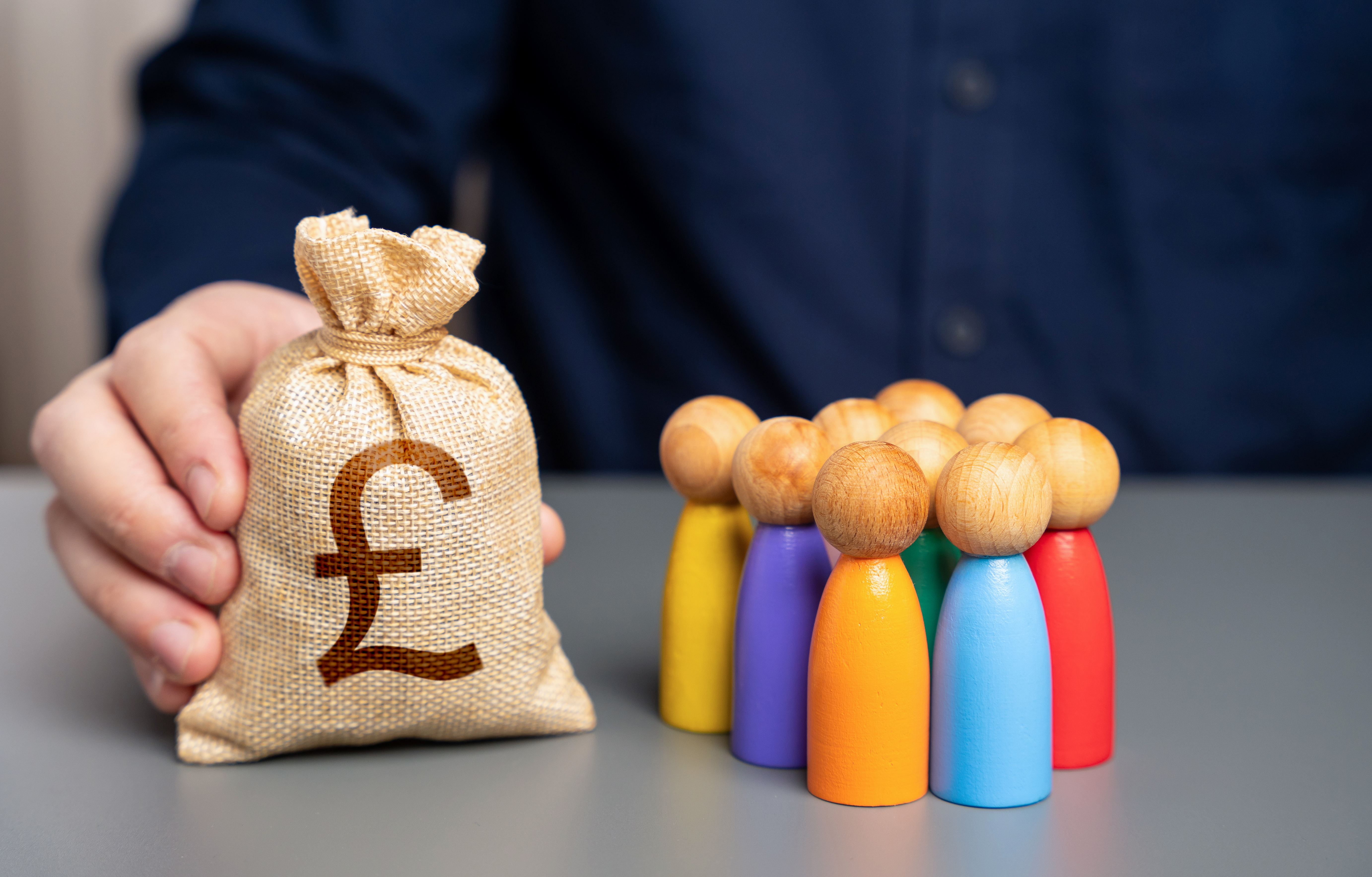 A photograph depicts a hand holding a small, beige burlap sack with a brown £10 coin visible. In the background, a person in a dark blue shirt is blurred. In the foreground, a row of colorfu