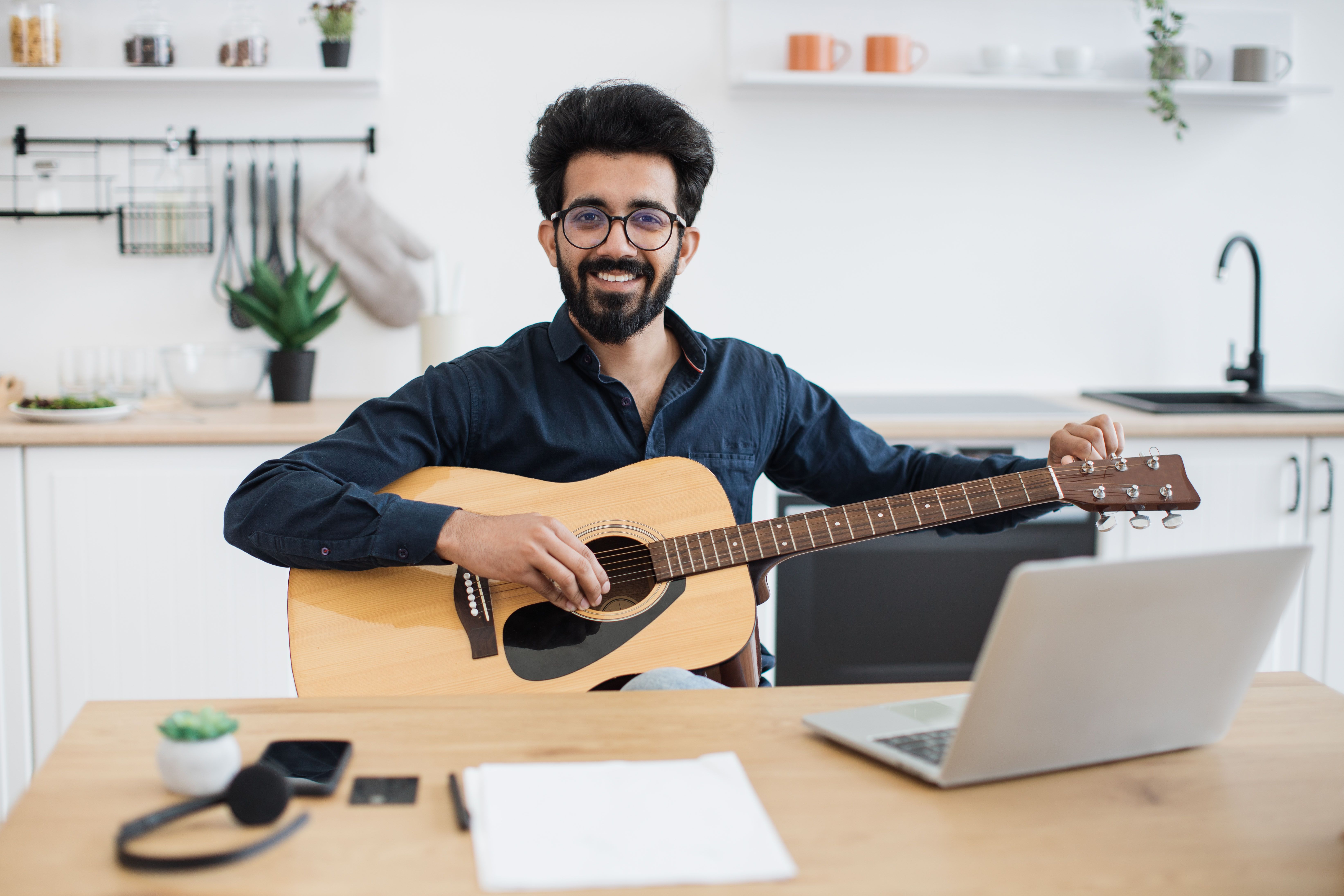 A photograph captures a smiling South Asian man with a full beard and curly black hair, wearing glasses and a dark blue shirt, playing a wooden acoustic guitar on a light wooden desk.