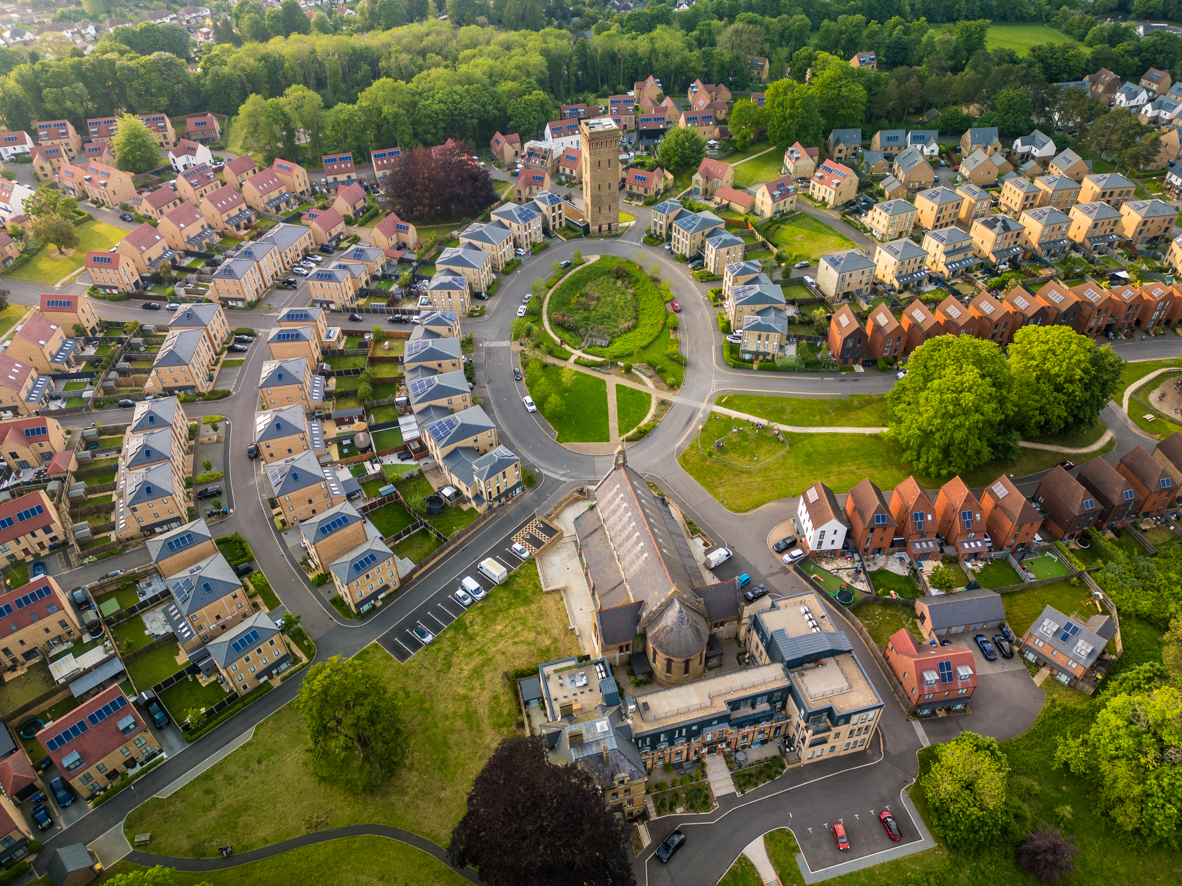 This is an aerial photograph of a modern suburban neighborhood featuring a central roundabout with a tall, stone church tower.