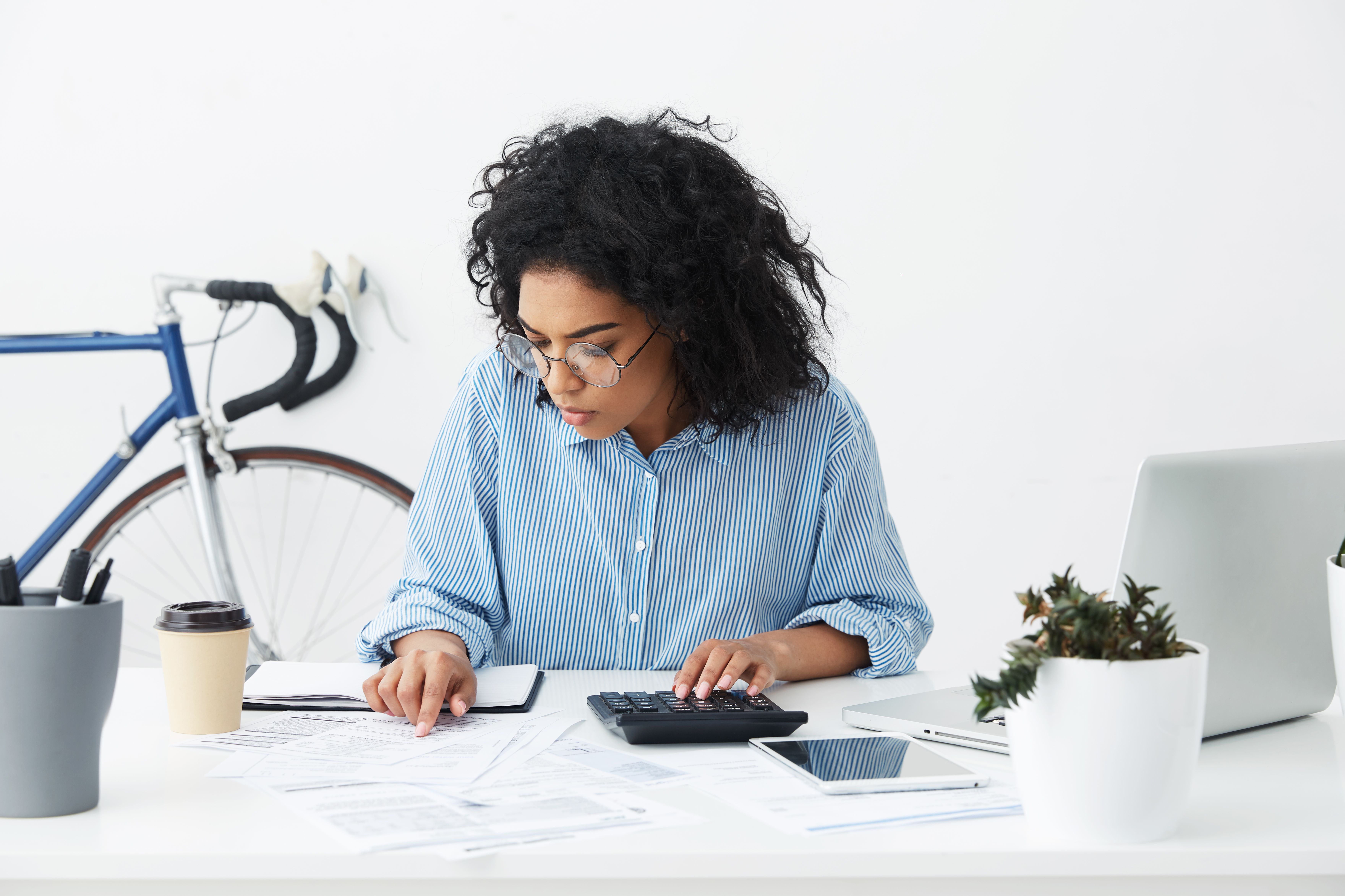 A photograph captures a curly-haired, light-skinned woman with glasses, wearing a blue and white checkered shirt, sitting at a white desk. She is focused on her documents and a calculator, with a laptop beside her.
