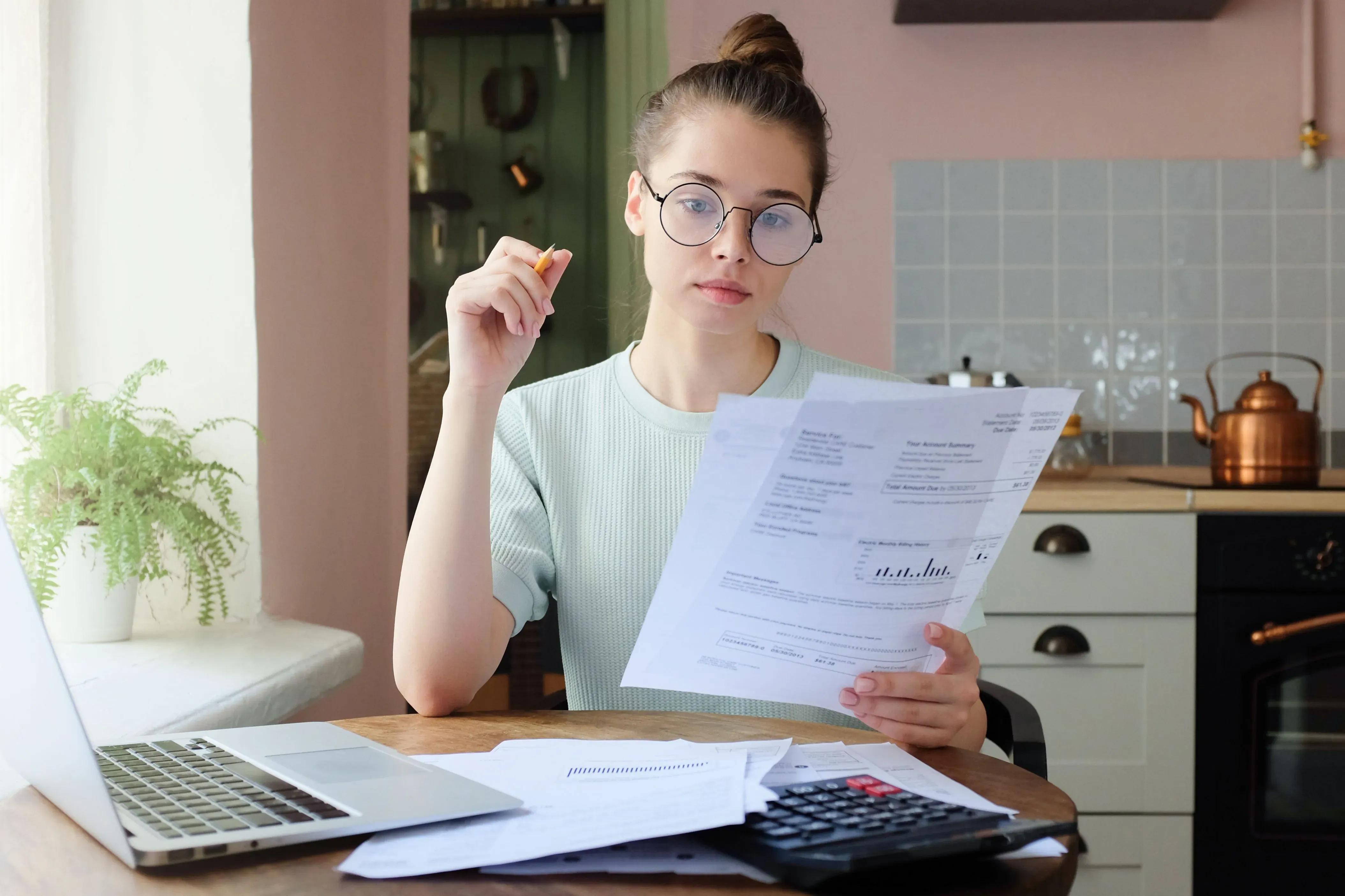 Photograph of a young woman with fair skin, light brown hair tied in a bun, and wearing round glasses, sitting at a wooden table in a kitchen. 