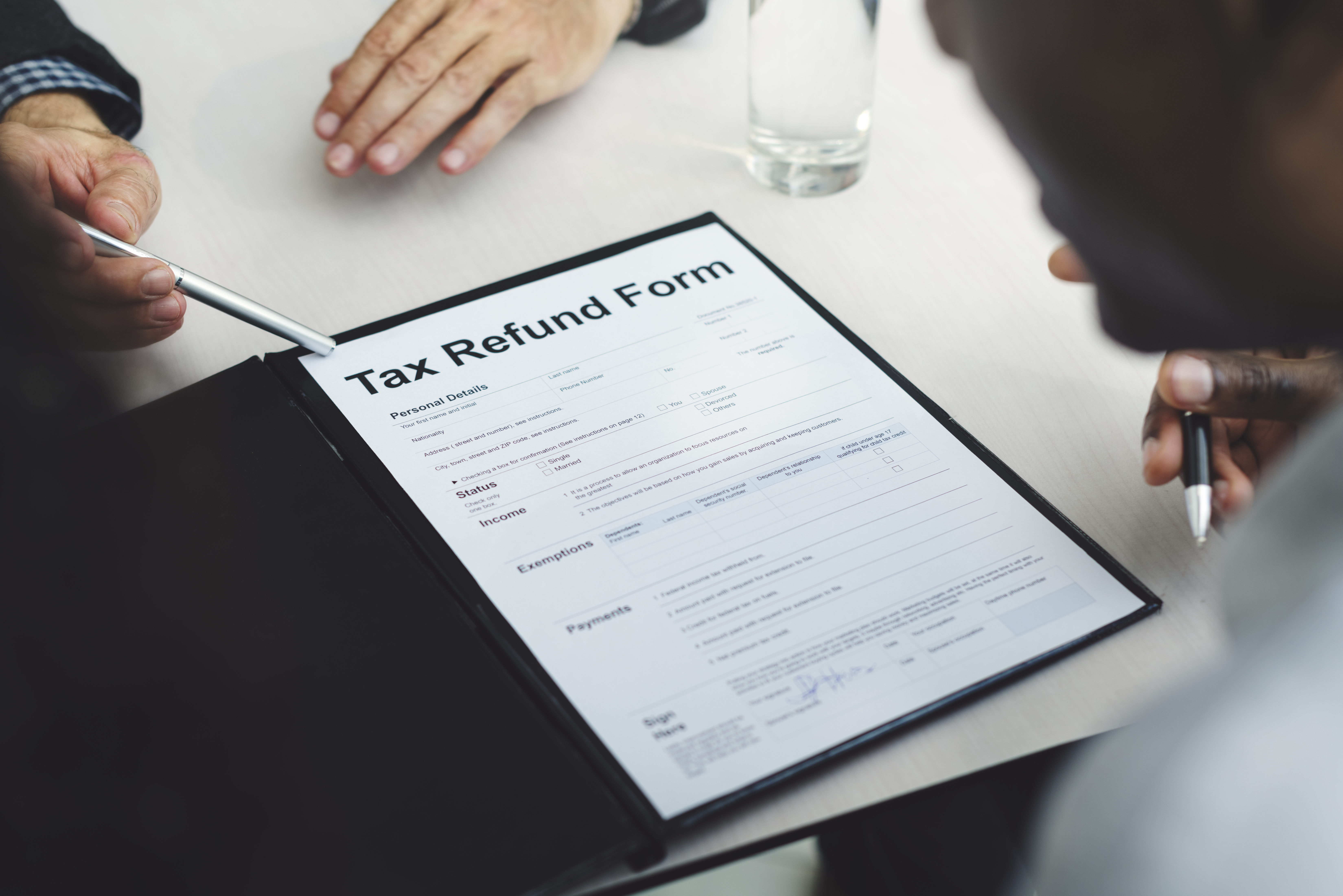 Photograph of a meeting scene with two individuals, one Black and one White, seated around a light-colored table. The Black person, wearing a dark suit, holds a pen, pointing to a "Tax Refund Form" document on a black clipboard.