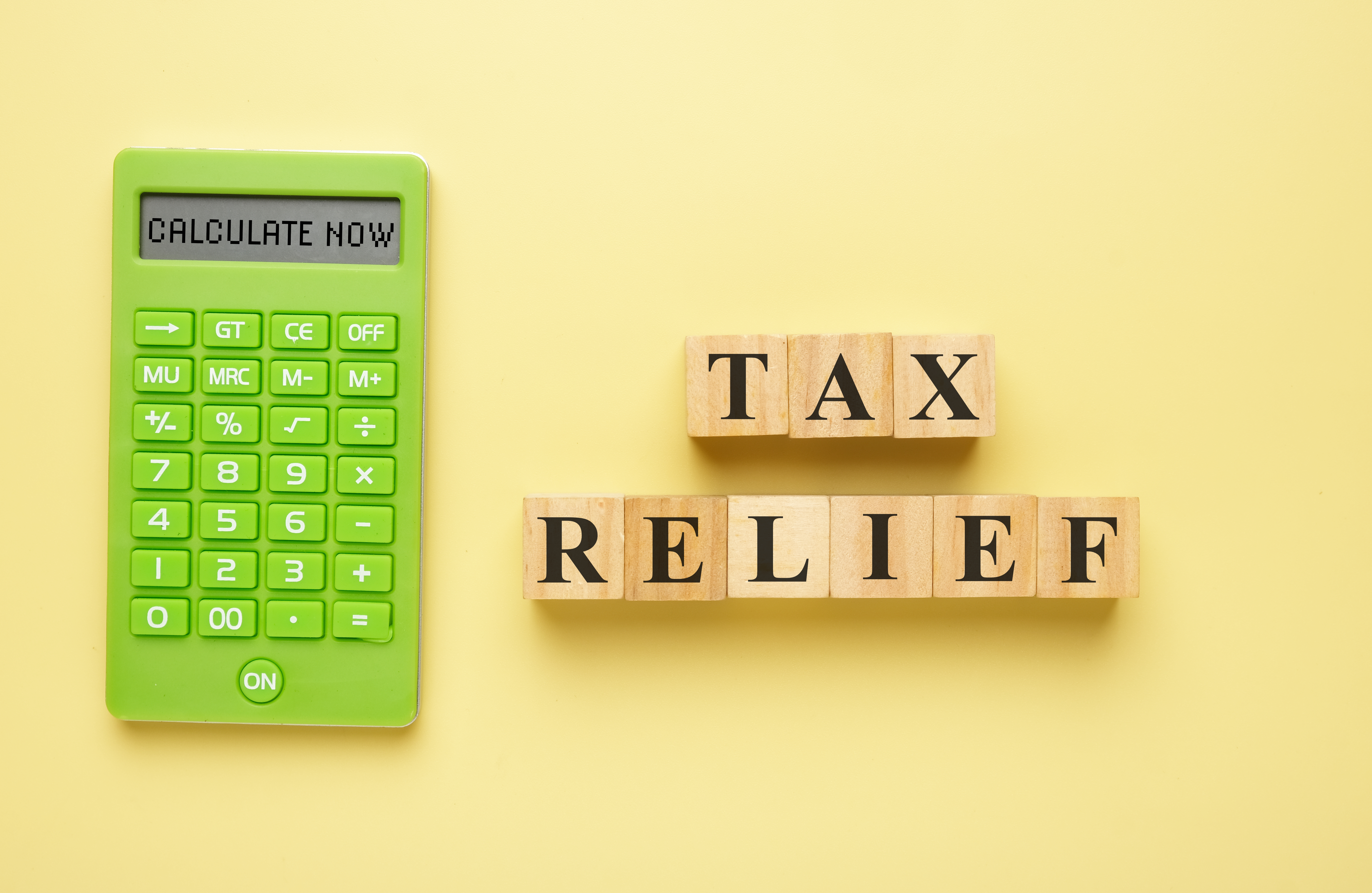 A photograph features a bright yellow background. On the left is a large, green calculator with a digital display reading "CALCULATE NOW." To the right, three wooden blocks are arranged horizontally, spelling out "TAX RELIEF" in black letters.