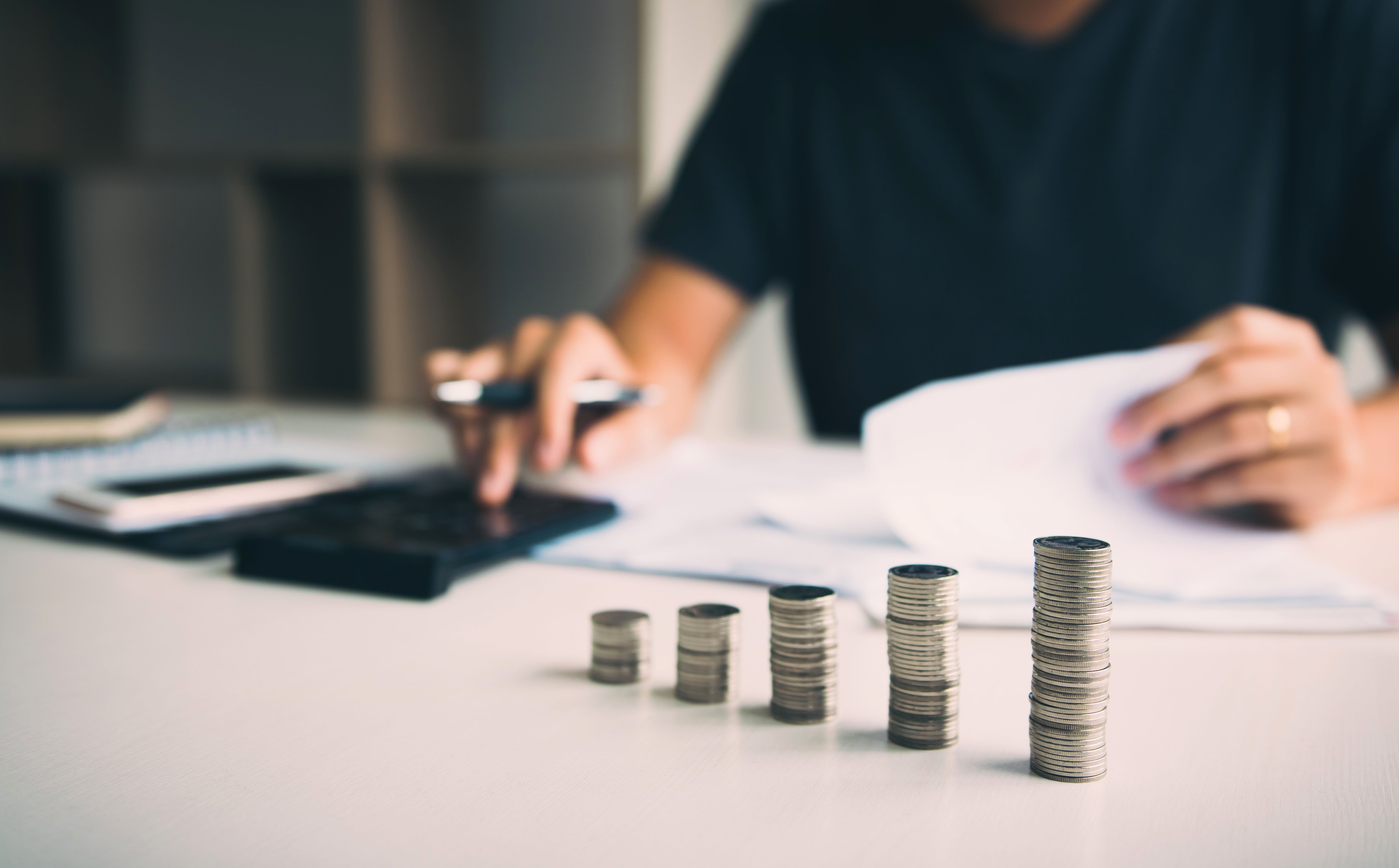 Photograph of a person, wearing a black T-shirt, seated at a white desk, focused on counting stacks of coins. 
