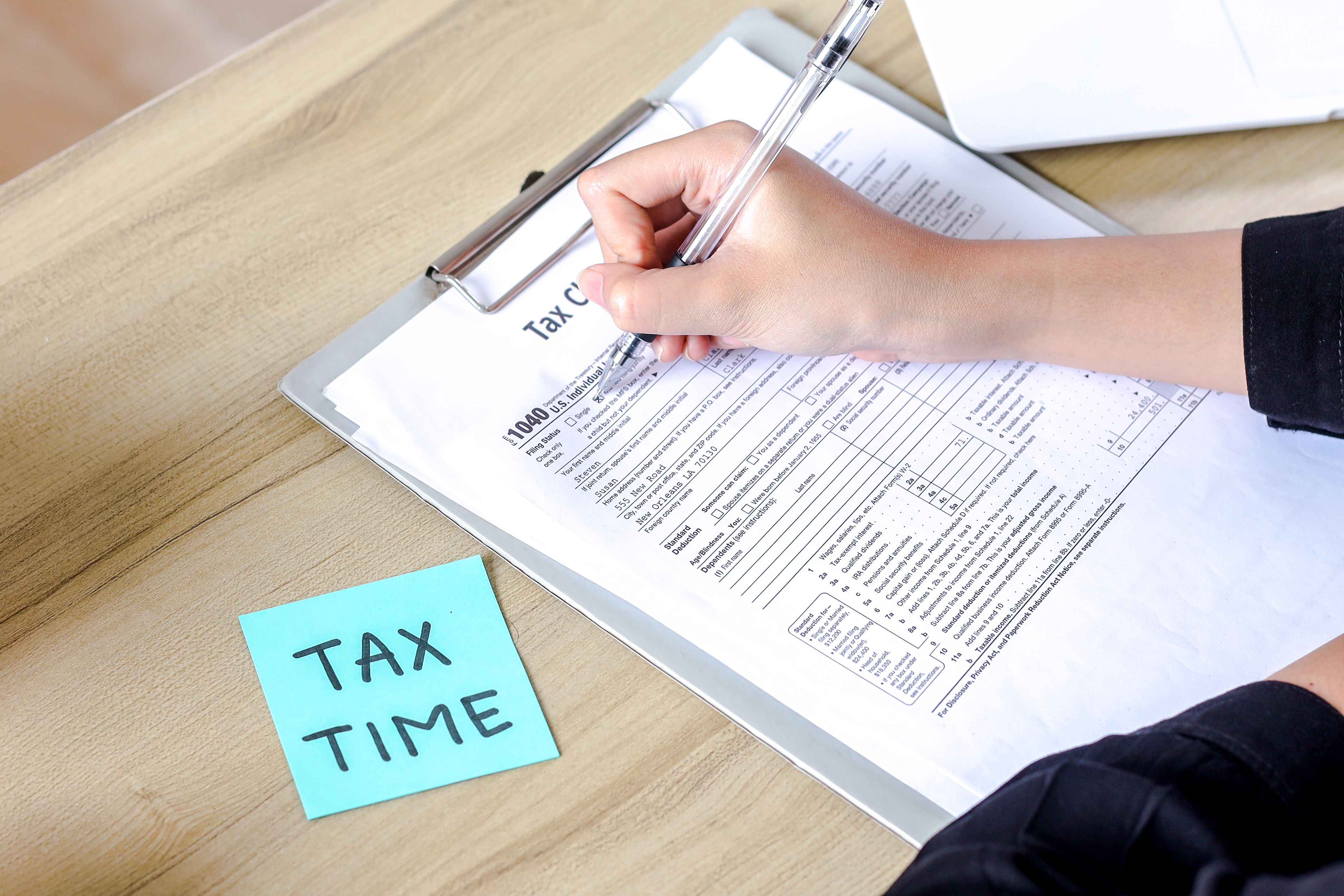 A photograph depicts a person with light skin writing on a clipboard with a pen.