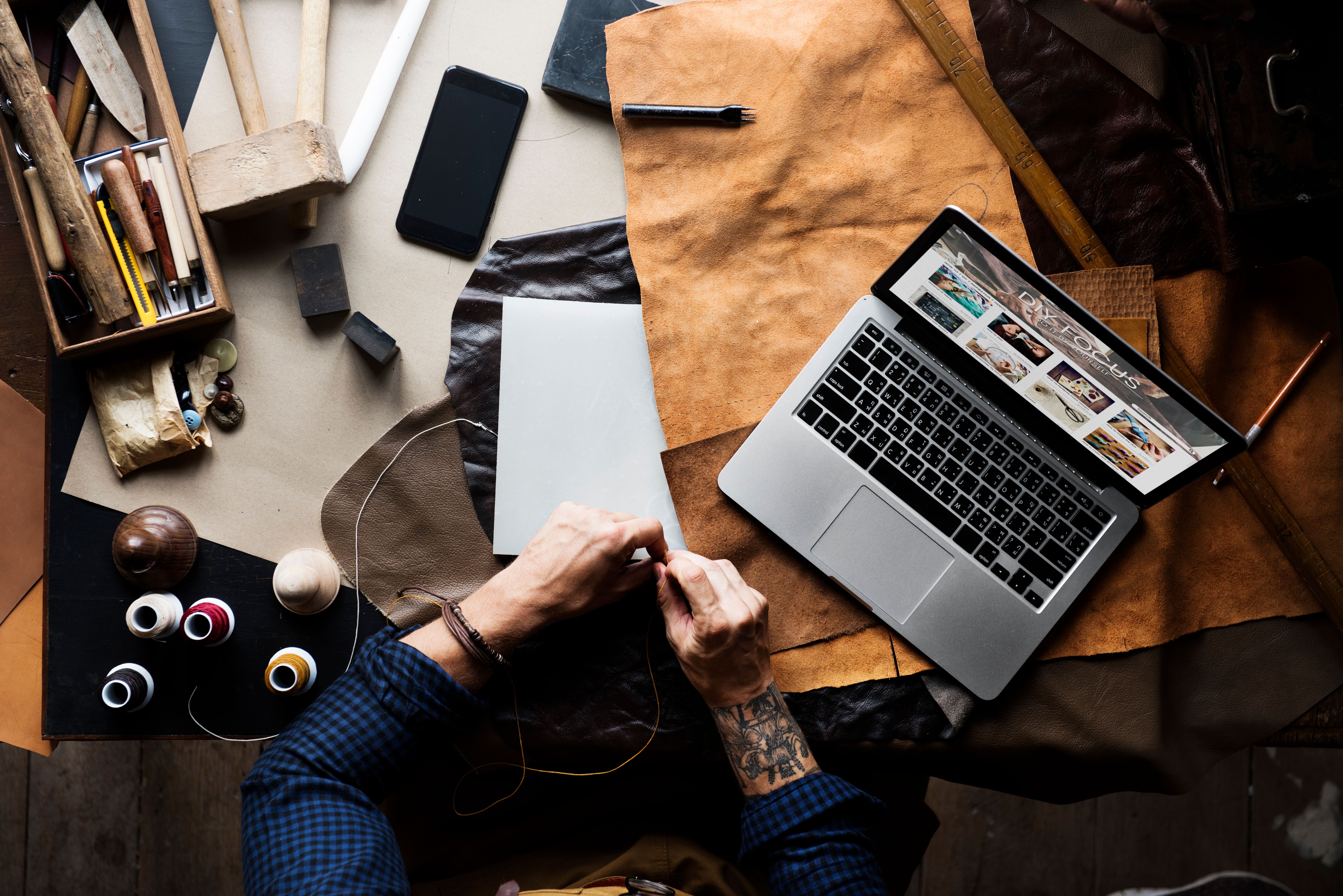 A photograph of a leather craftsman's workspace, featuring a person with tattoos in a blue plaid shirt, sewing leather. 
