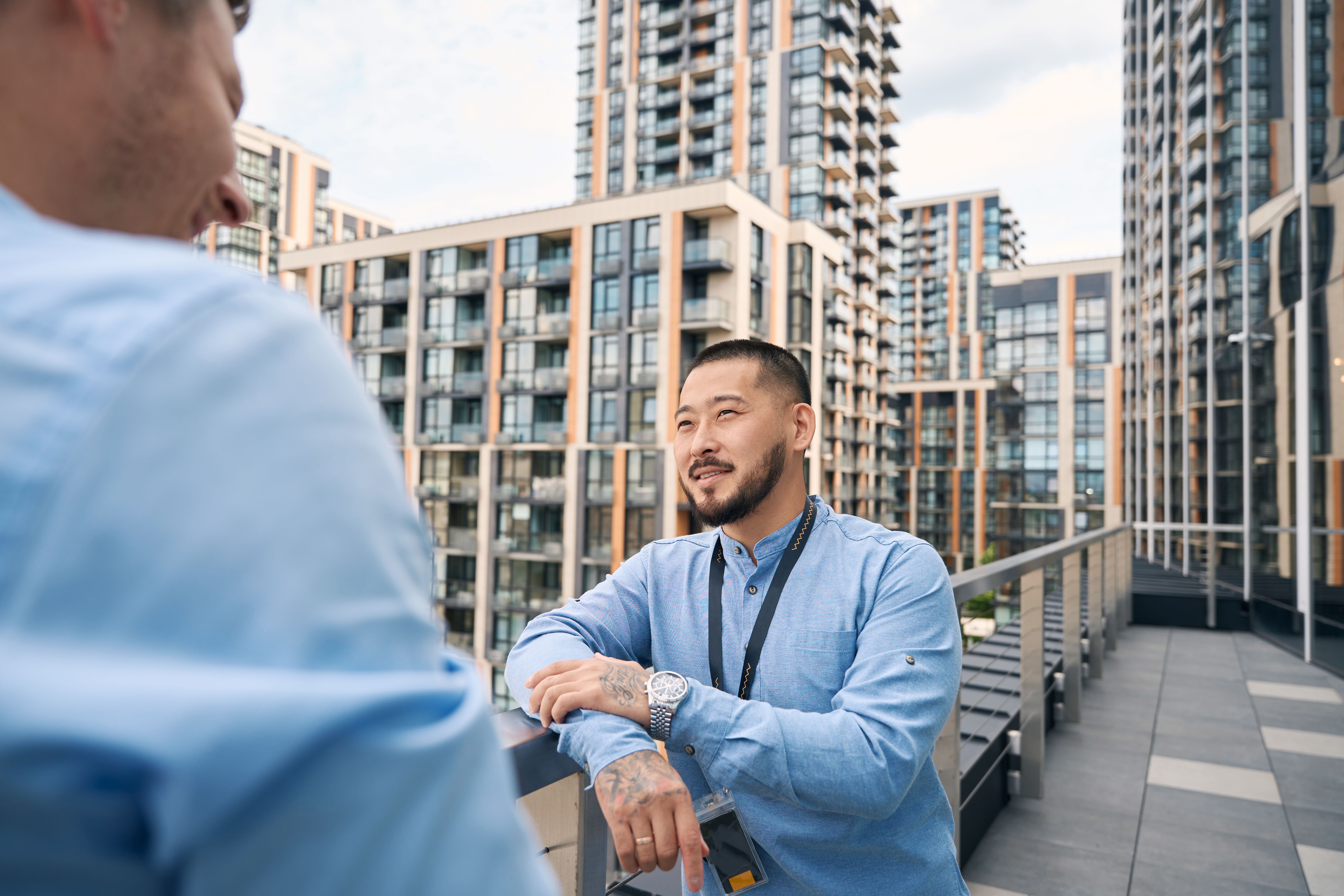 A photograph shows a bearded man with tattoos on his arms, wearing a light blue button-up shirt and a lanyard with a badge, leaning against a balcony railing. 