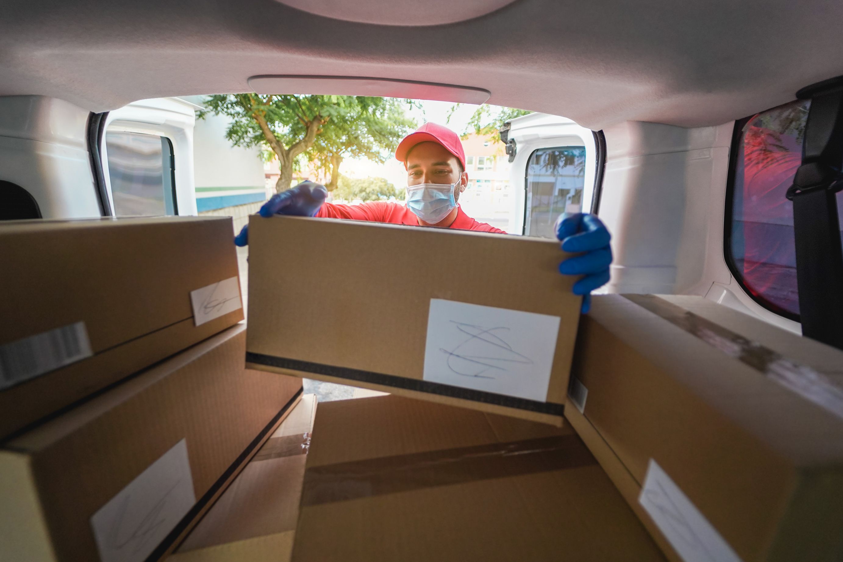 A delivery worker, wearing a red cap, blue gloves, and a face mask, standing inside a white van.
