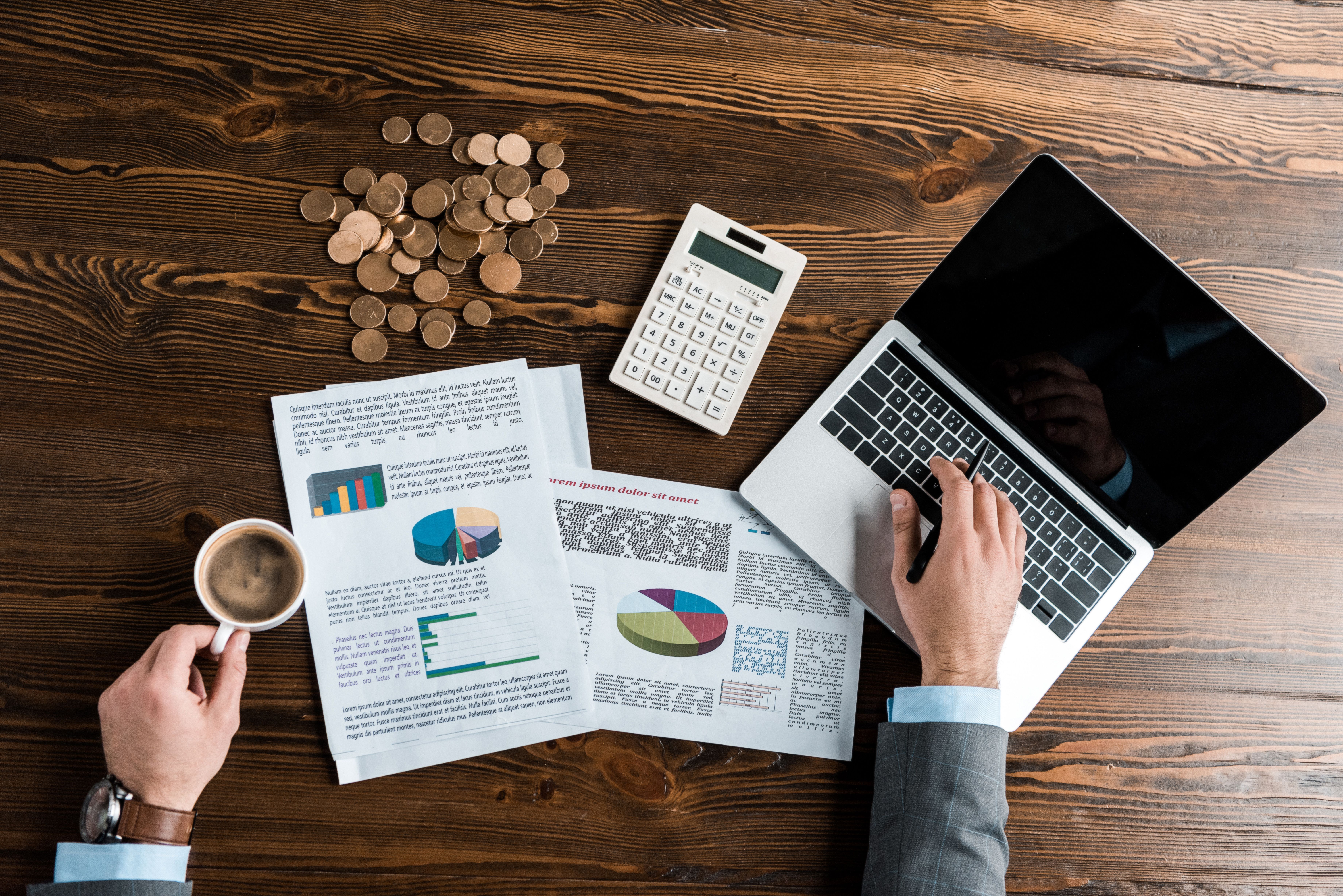 This is a photograph of a top-down view of a wooden desk. A hand in a gray suit jacket holds a white coffee cup with a brown liquid. A silver laptop is open, showing a black screen.