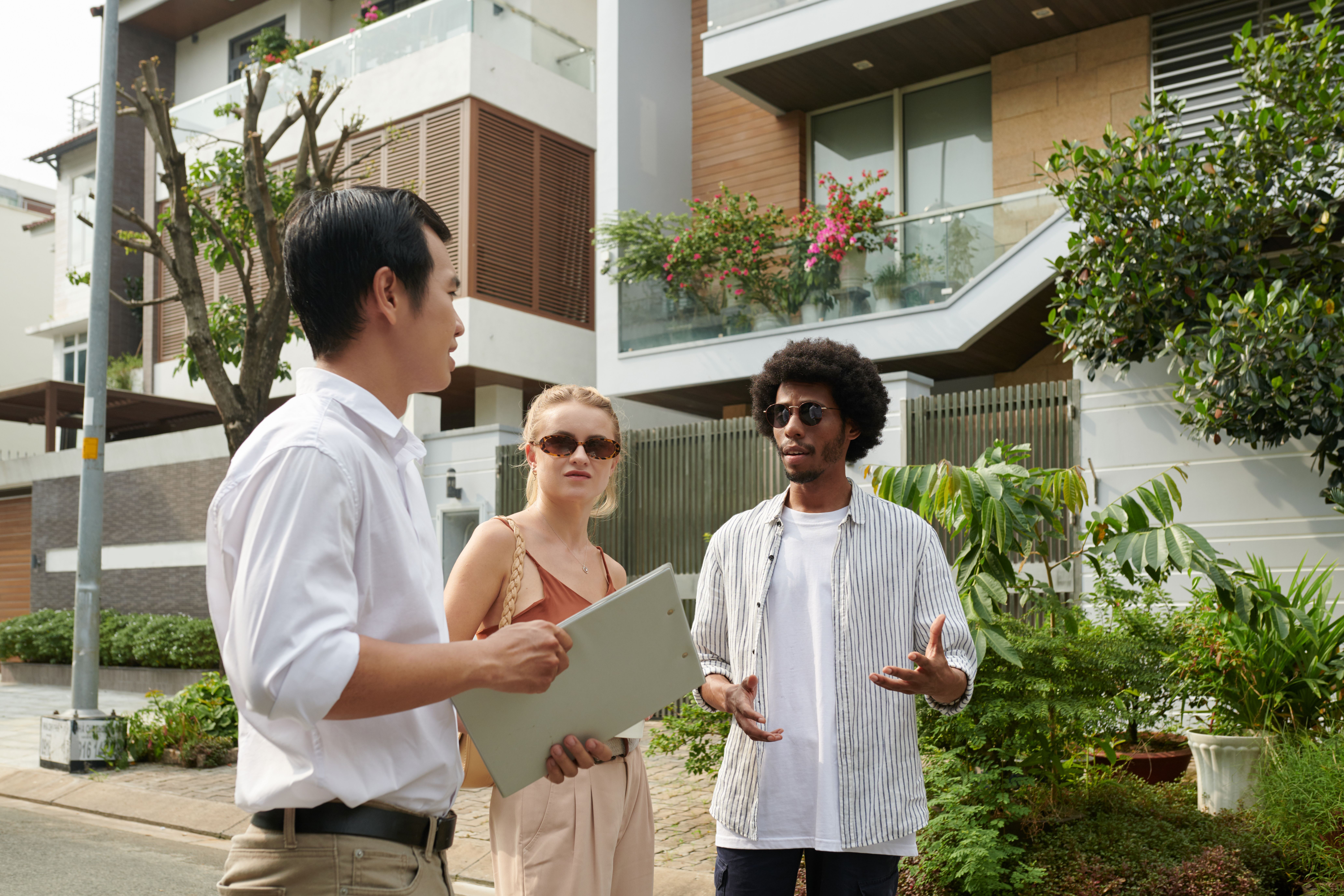 A photograph captures three individuals standing on a suburban street. A man with short black hair in a white shirt and beige pants holds a clipboard.