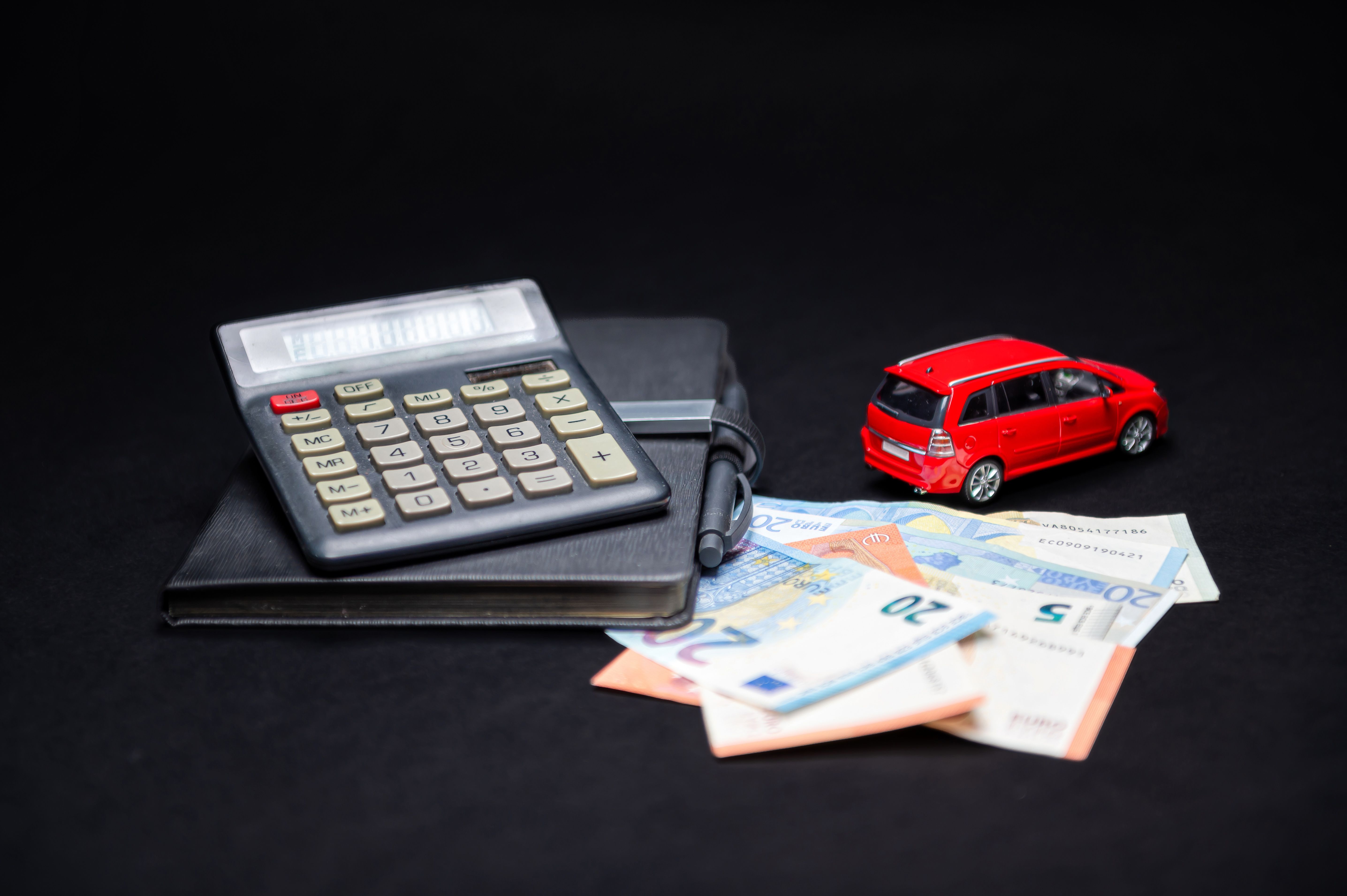 A photograph depicts a black calculator with a clear display, placed atop a closed black leather notebook, resting on a black fabric surface. Scattered around the notebook are various European currencies, including euros, and a small red toy car, position
