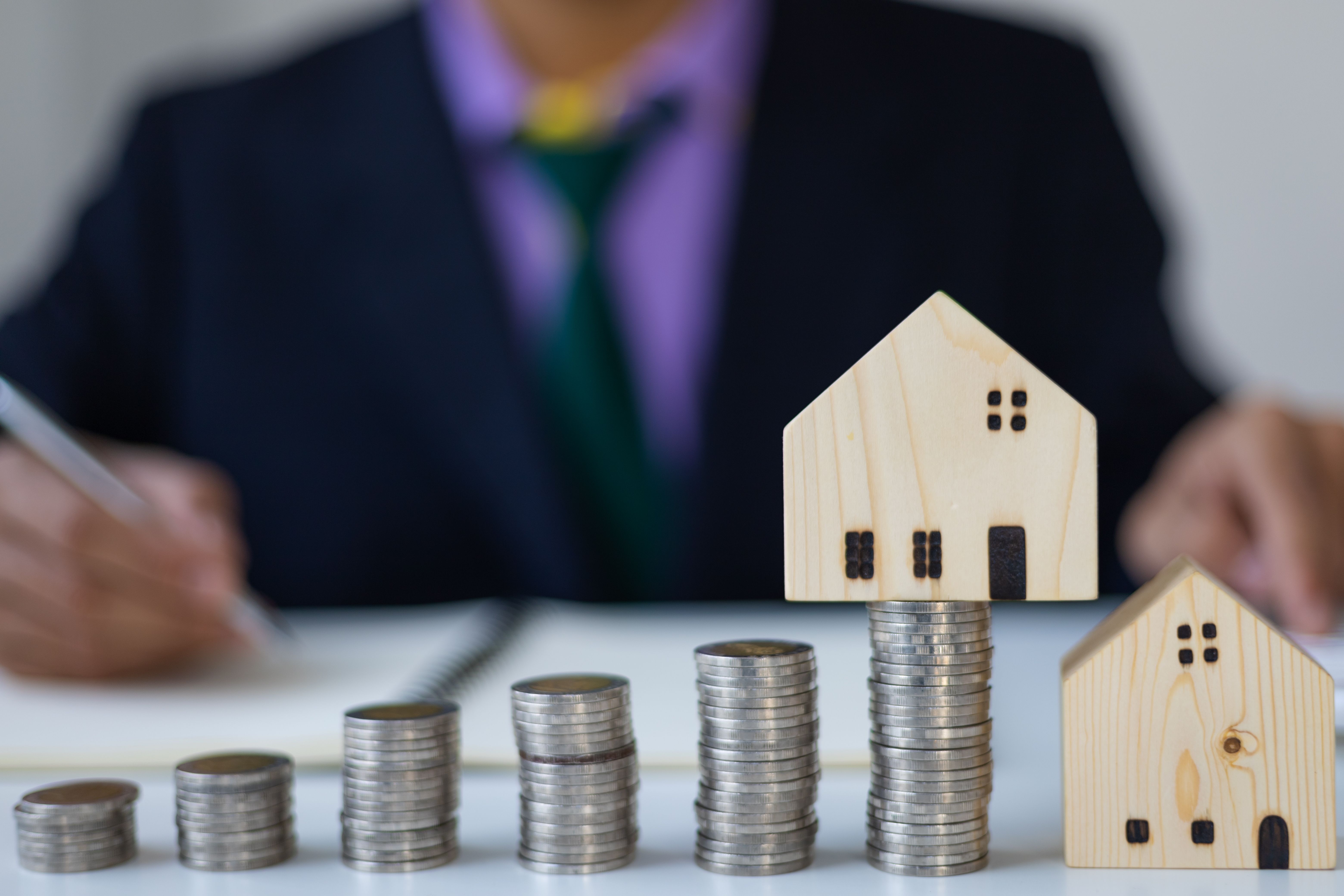 A photograph shows a person in a dark suit and purple shirt, blurred in the background, writing with a pen on a notepad. In the foreground, a wooden house model with a stack of coins beneath it, symbolizing savings or investment. 