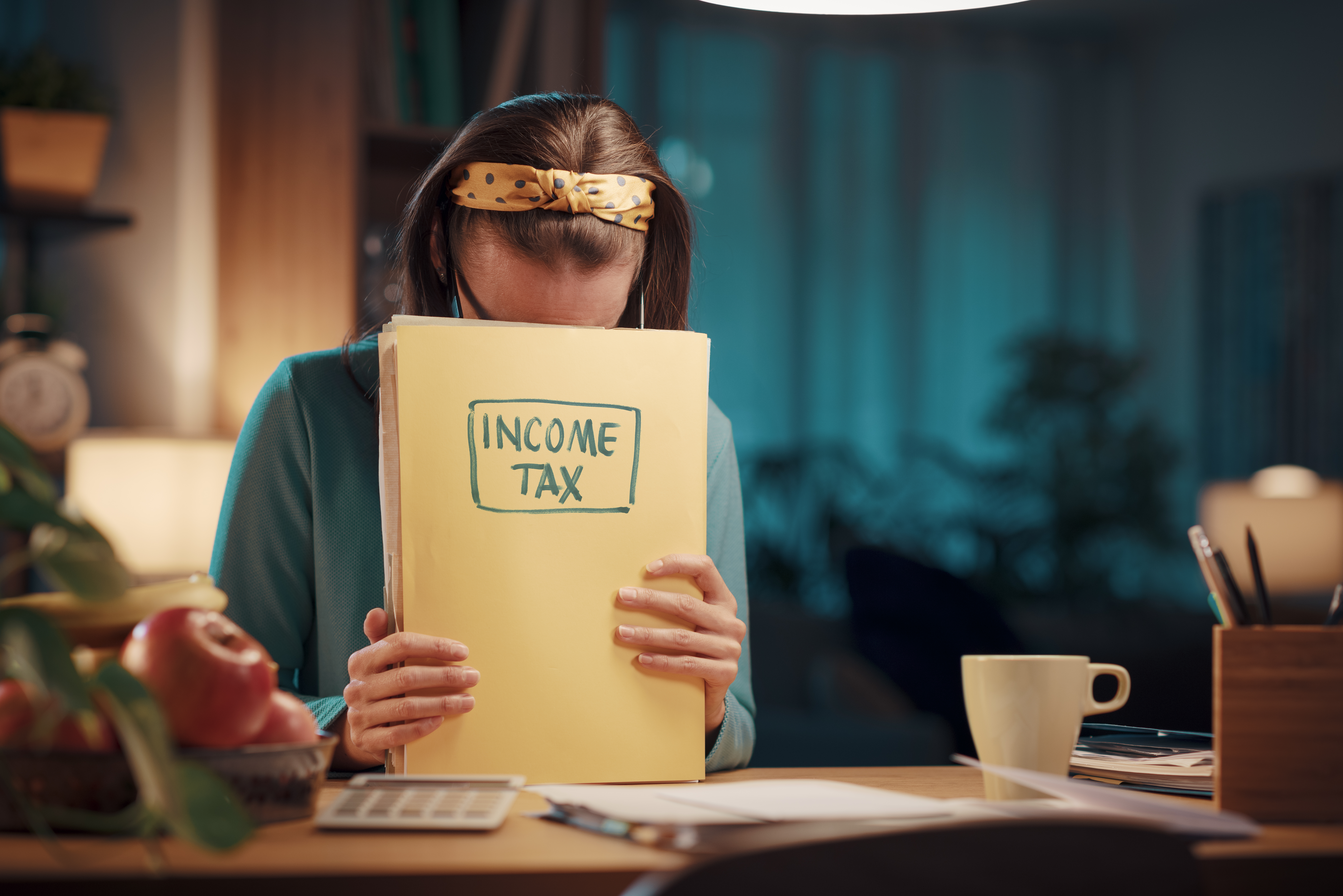 Woman with long brown hair, wearing a teal sweater and a yellow polka-dot headband, sits at a cluttered desk. She is reading a yellow notebook labeled "INCOME TAX."