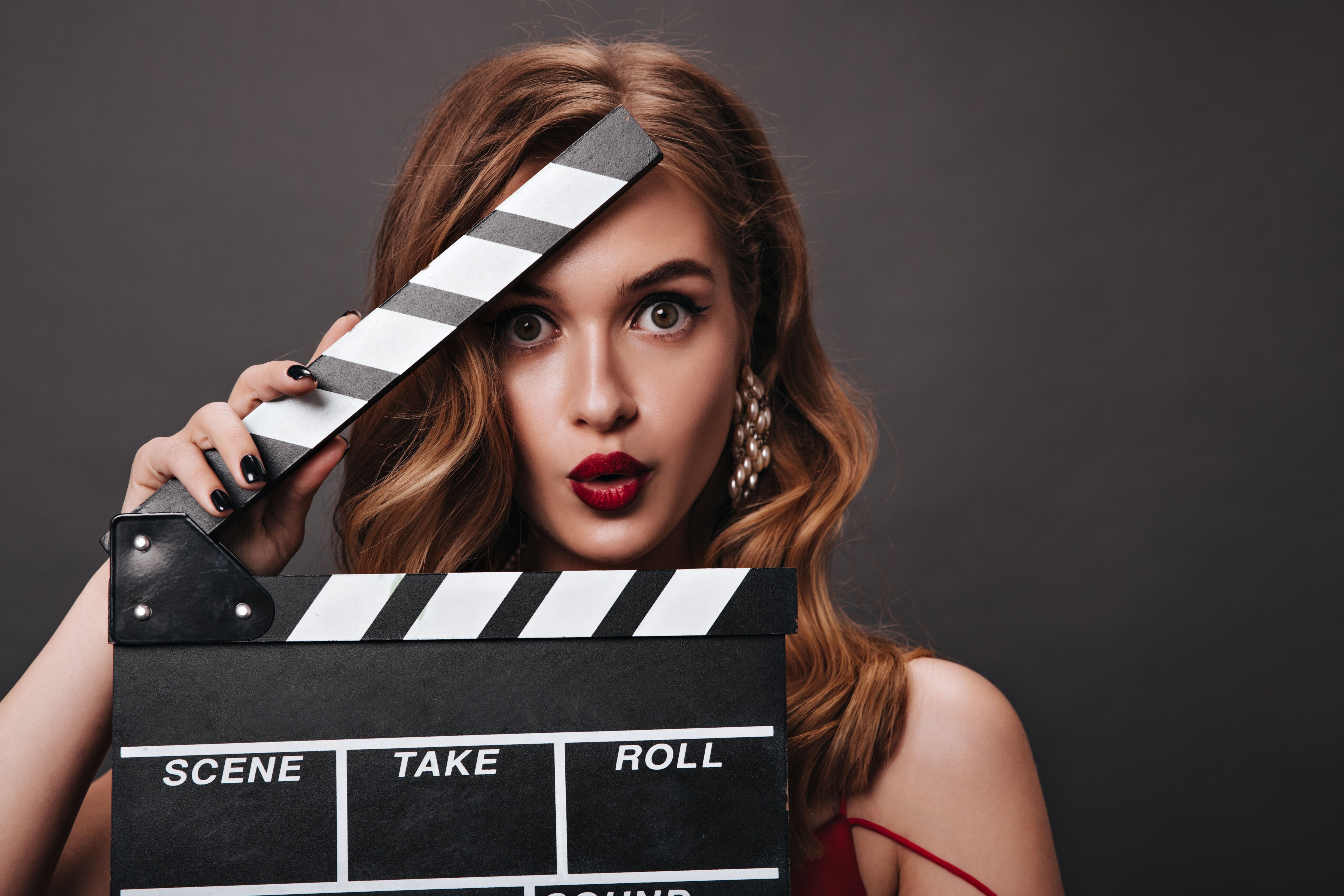 A photograph featuring a young Caucasian woman with wavy, light brown hair, wearing bold red lipstick and pearl earrings, holding a black and white film clapperboard in front of her face.