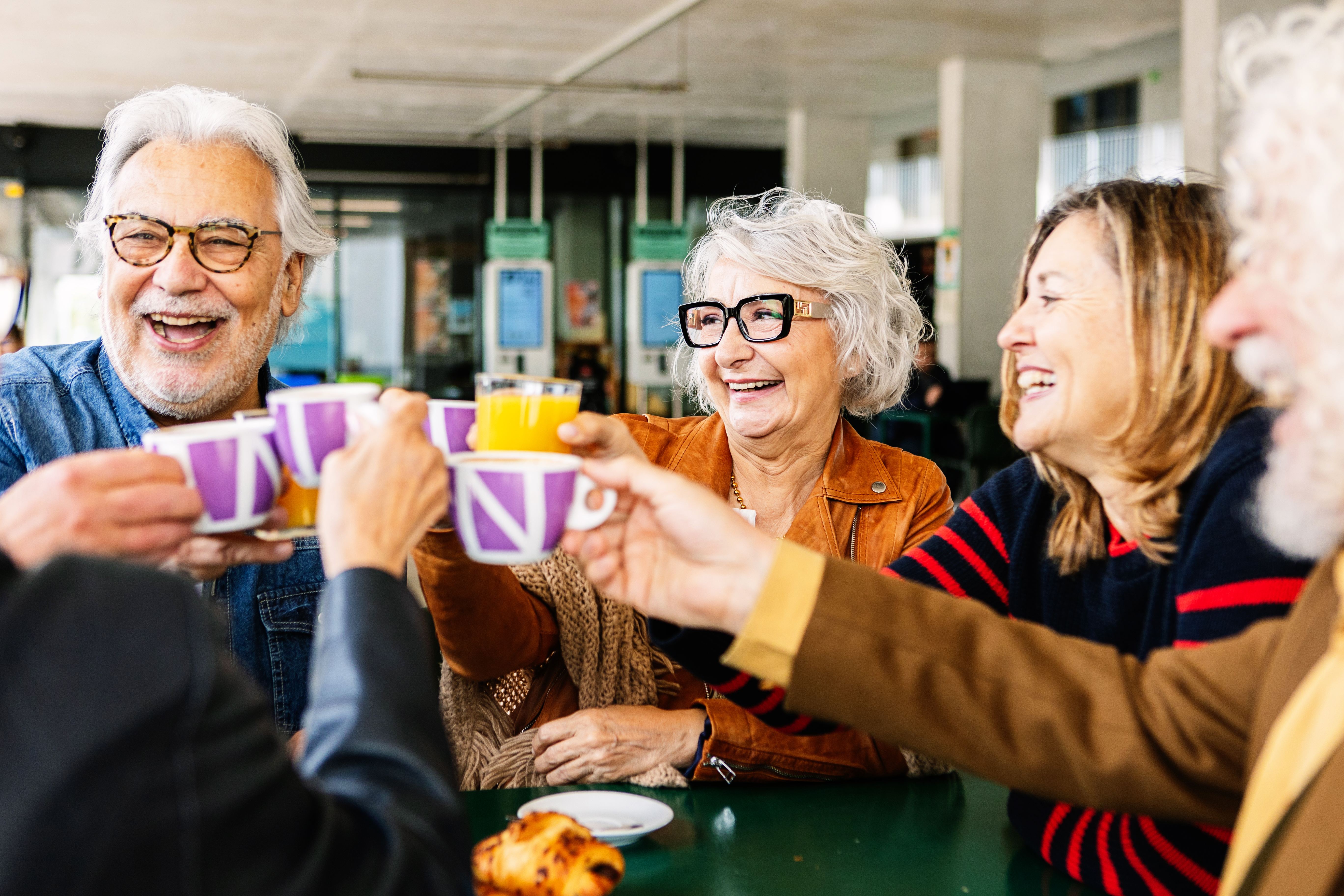 A vibrant photograph captures four elderly individuals, two men and two women, joyfully toasting with colorful mugs at a casual café.