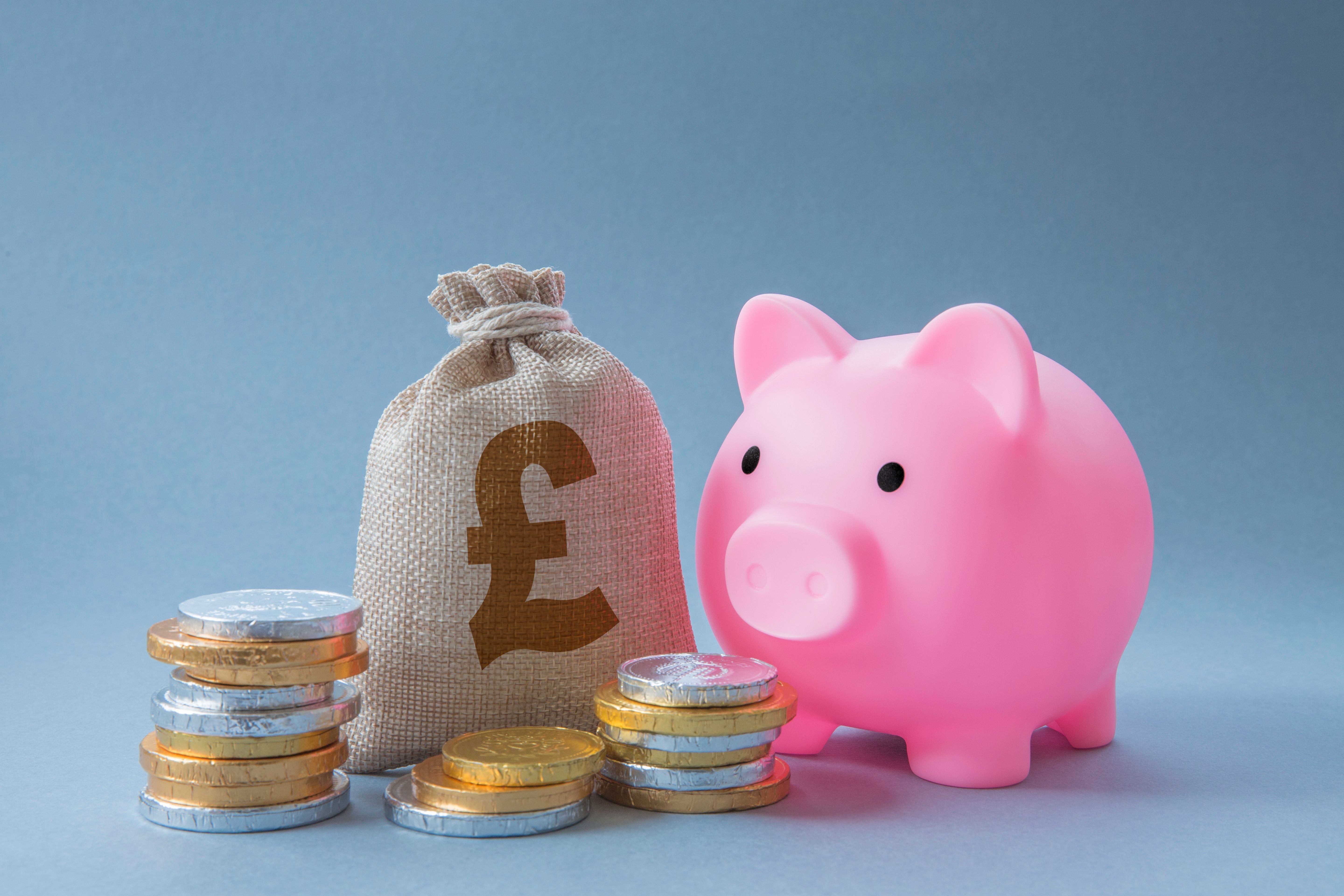 Photograph of a minimalist setting featuring a pink plastic piggy bank with a smiling face and black eyes on the right. To the left, a beige fabric money bag with a visible brown £10 coin inside
