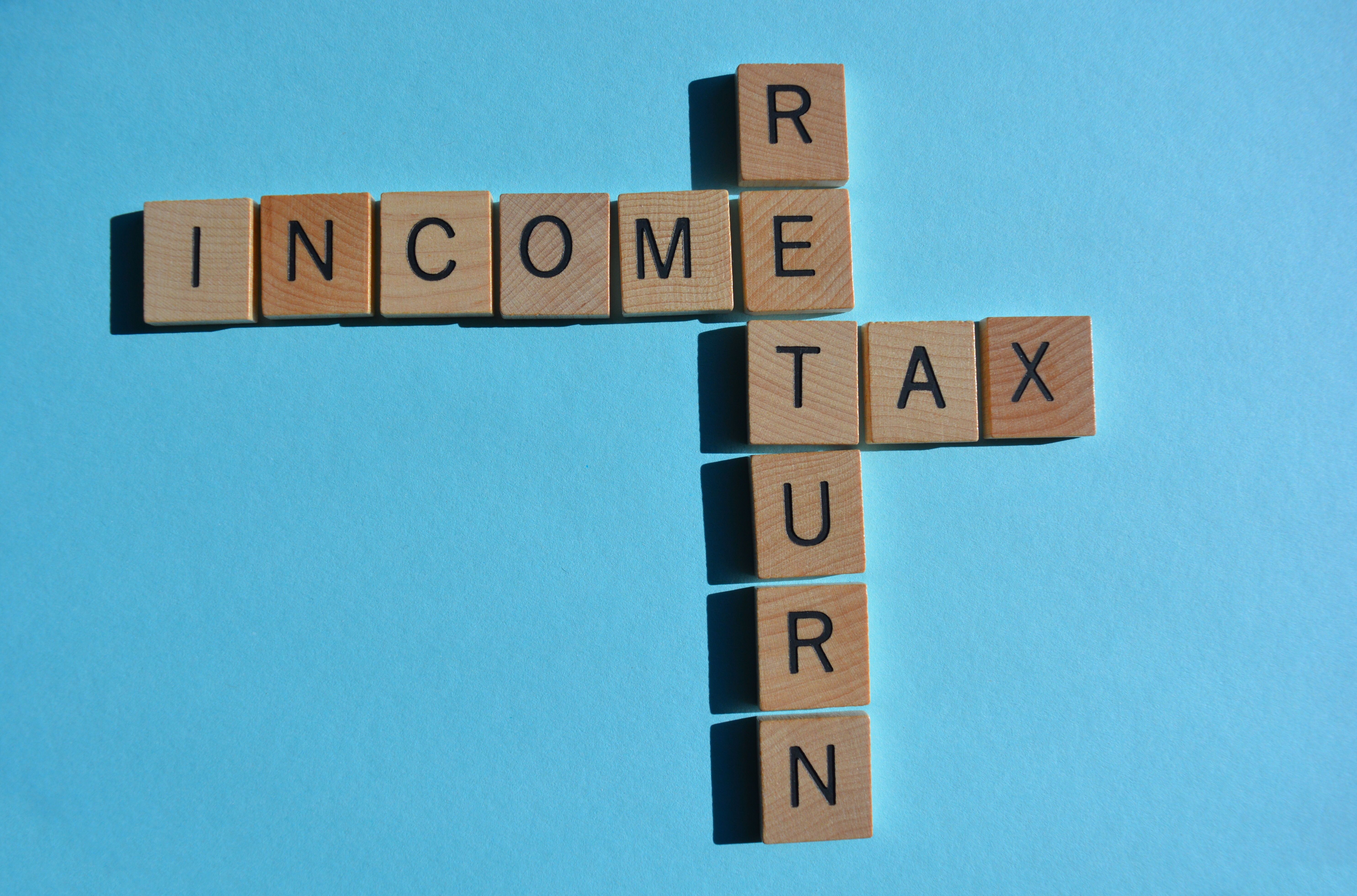 This photograph depicts a wooden Scrabble board with the letters "INCOME TAX RETURN" arranged in a cross pattern on a bright blue background. 