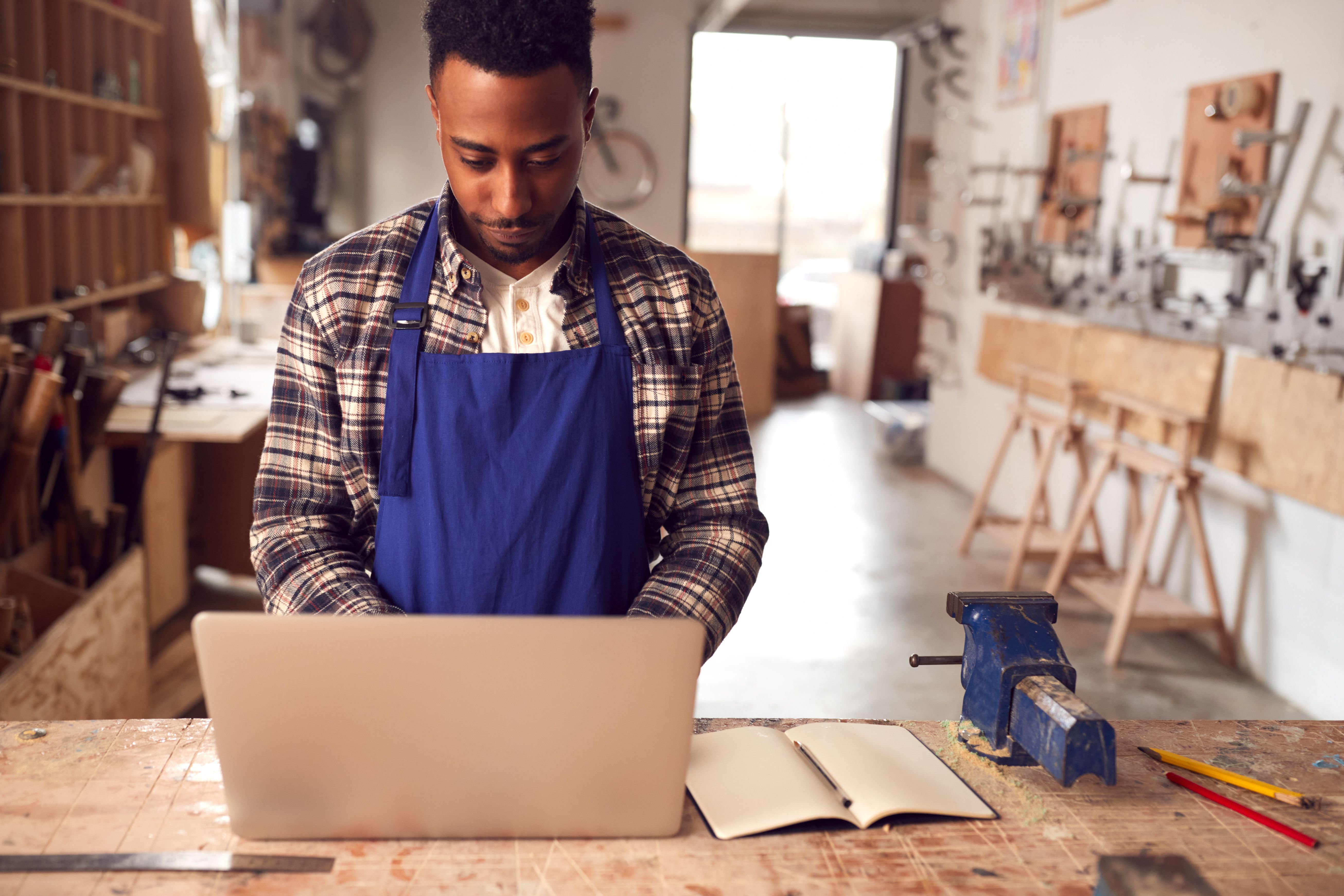 A photograph captures a young African-American man with a short, curly afro and beard, wearing a blue apron over a black and white plaid shirt, focused on his open laptop on a cluttered workshop table. 