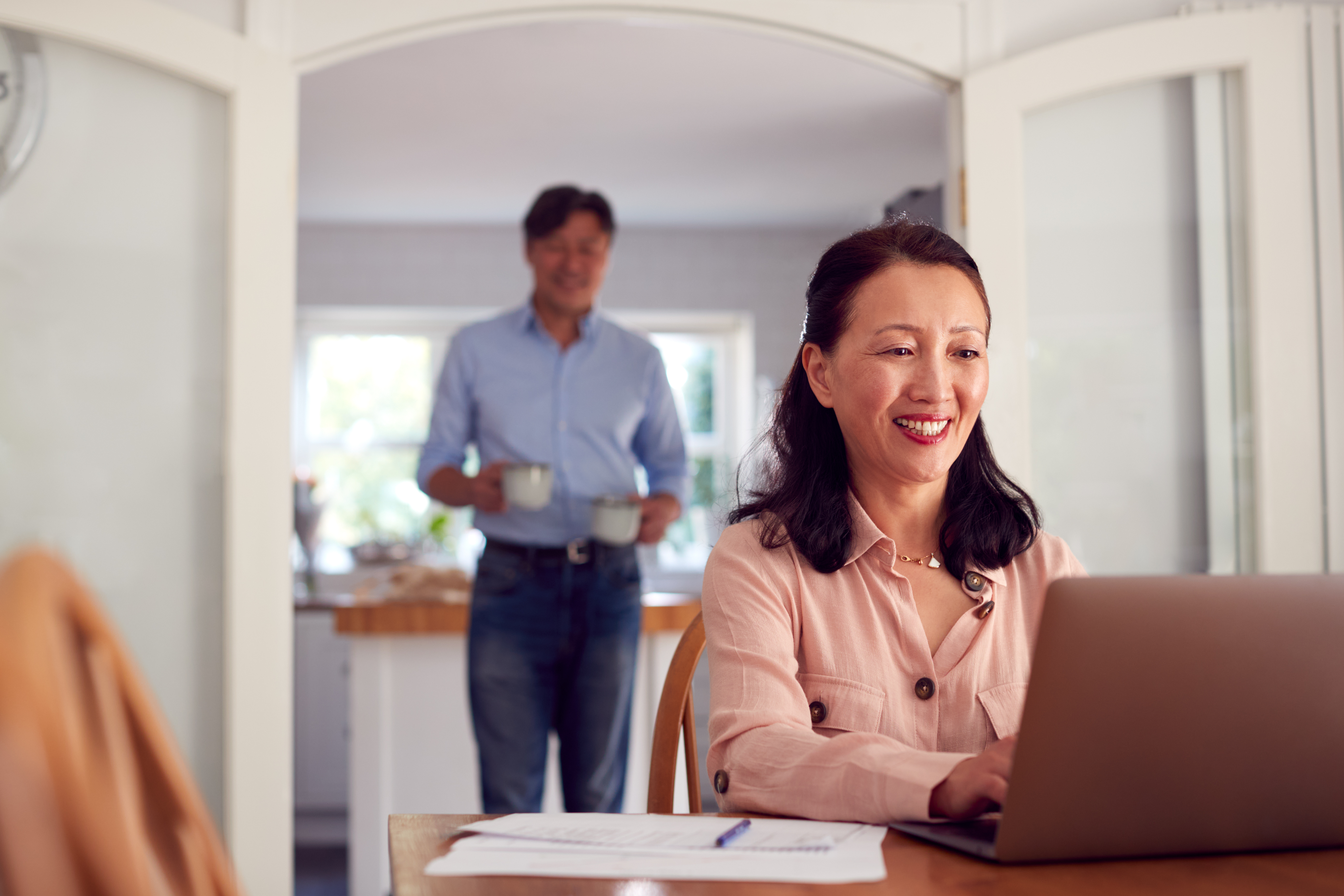 A smiling Asian woman with long black hair, wearing a light pink button-up shirt, sits at a wooden table with a laptop, in a bright, modern kitchen. 