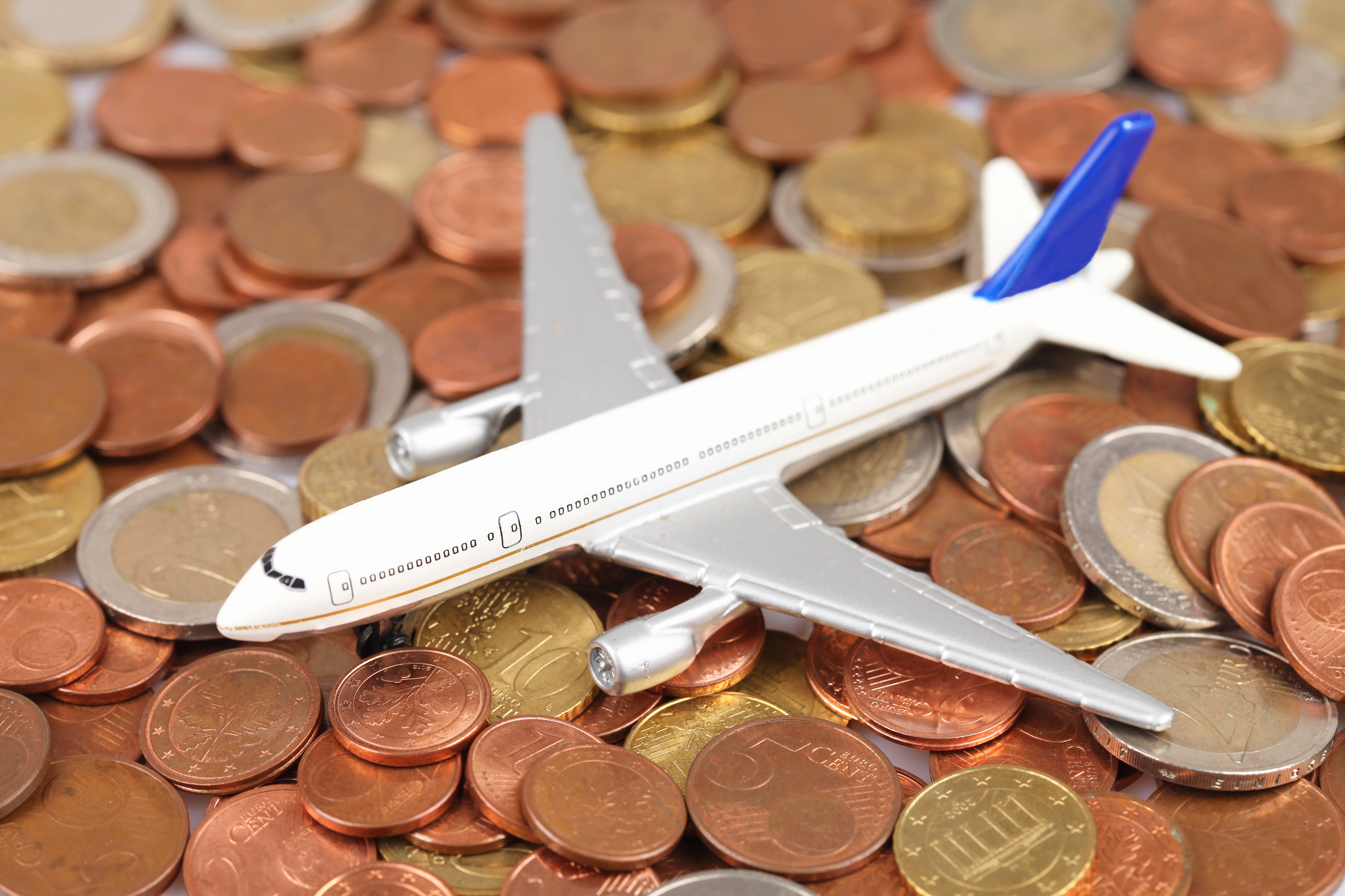 This is a photograph of a small, silver model airplane with a blue tail fin, resting on a pile of variously sized copper, bronze, and gold-colored coins.