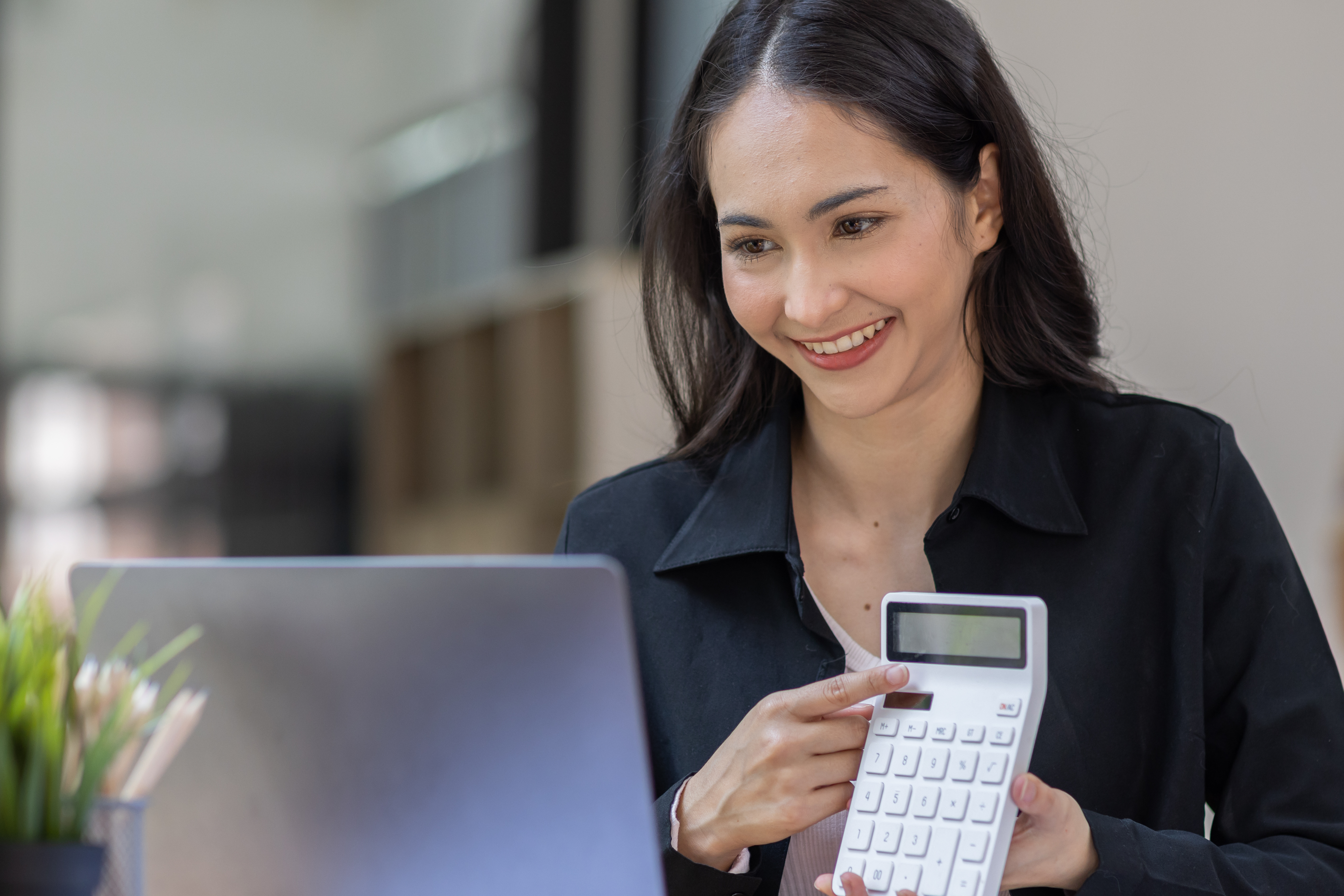 A photograph of a smiling, young woman with medium-length dark hair, wearing a black blazer. She is holding a white calculator and looking at a laptop screen in front of her. 