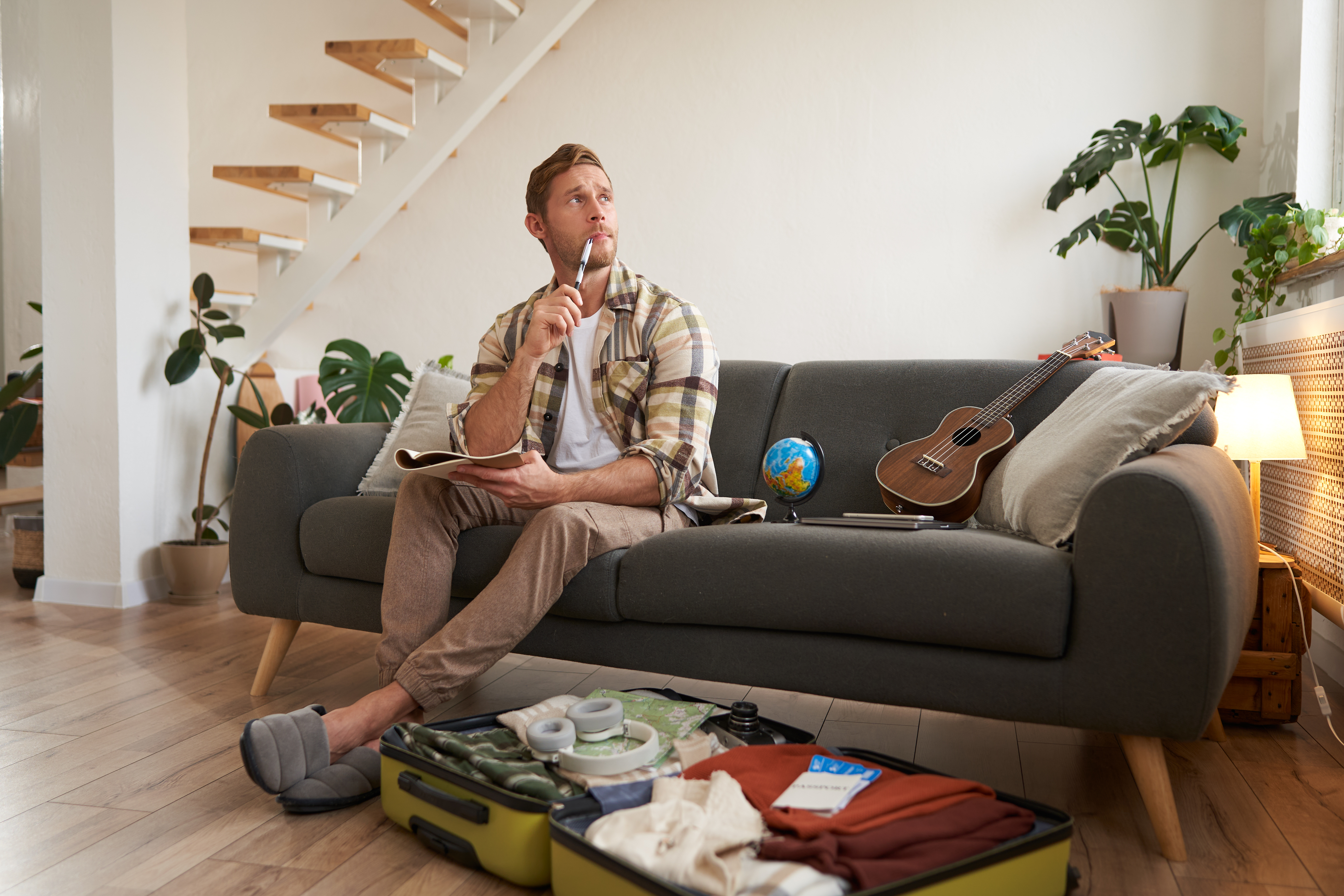 A photograph of a young man with short brown hair, wearing a plaid shirt and beige pants, sitting on a gray sofa in a modern living room. He holds a globe and a notebook, contemplating. A guitar rests beside him, and luggage is open on the floor. 