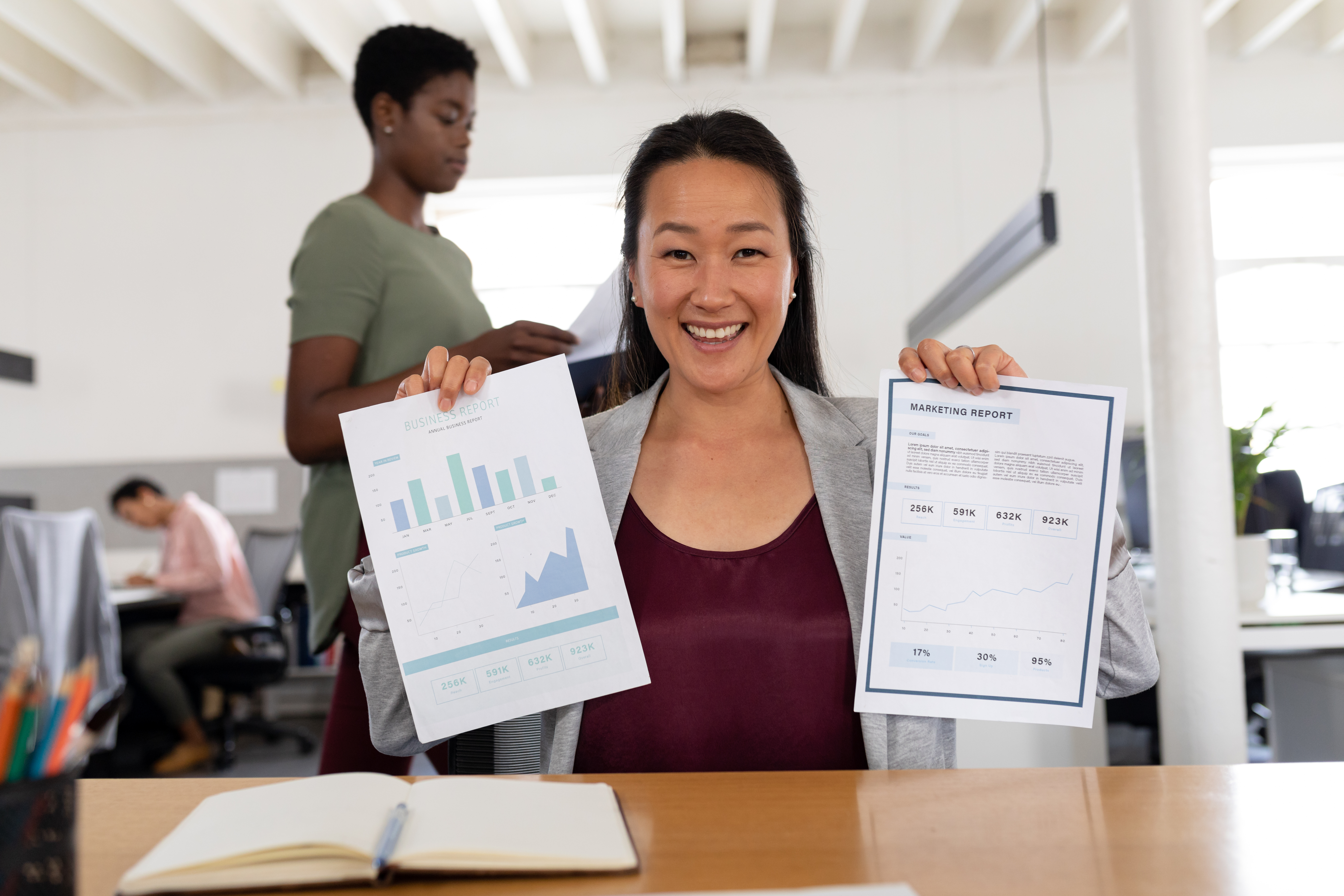 The photograph depicts a smiling Asian woman with long black hair, wearing a grey blazer over a burgundy top, holding two documents. The documents show a bar chart and a graph