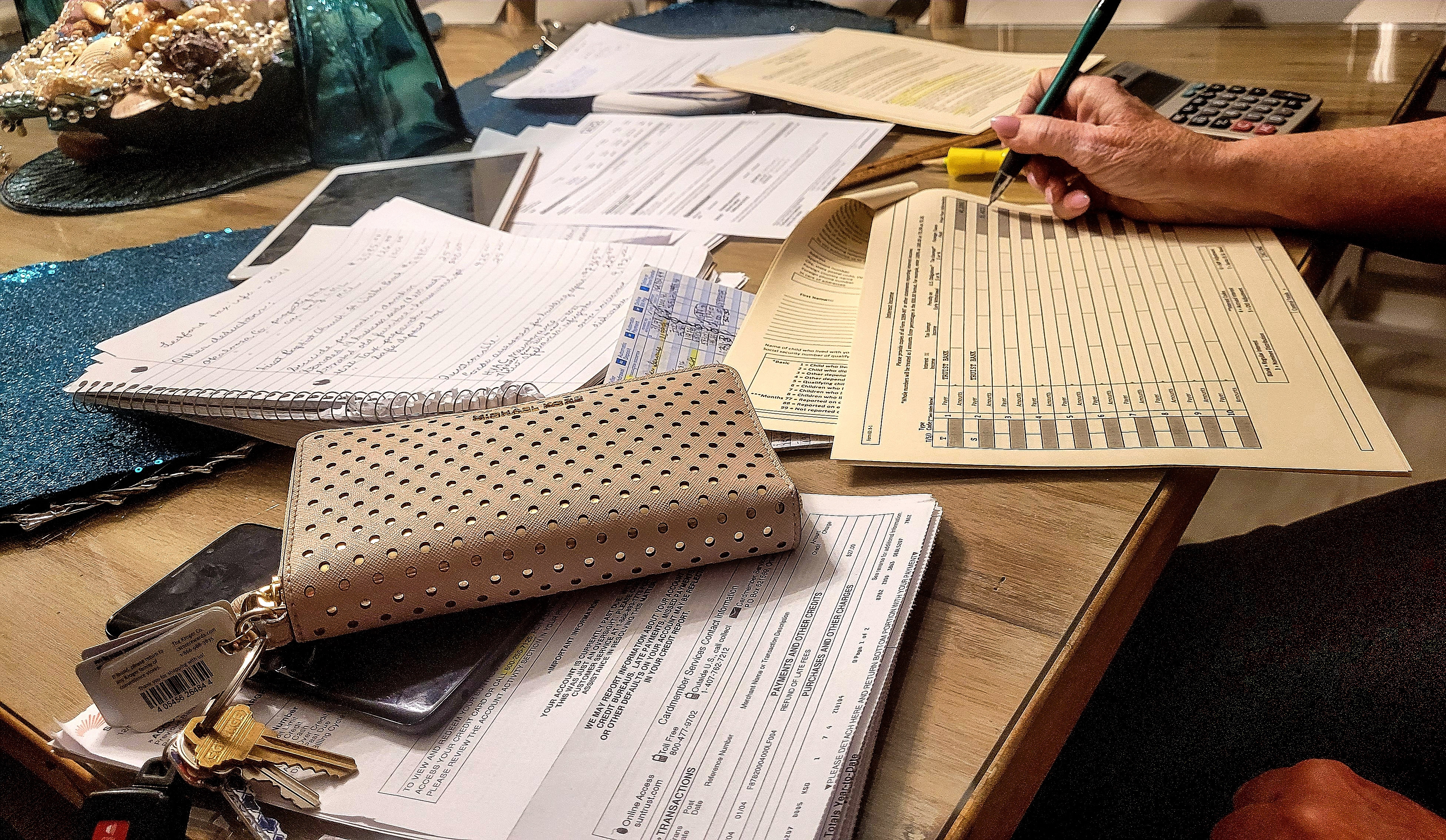 This photograph depicts a cluttered office desk with a hand holding a yellow pen, writing on a beige document with black text.