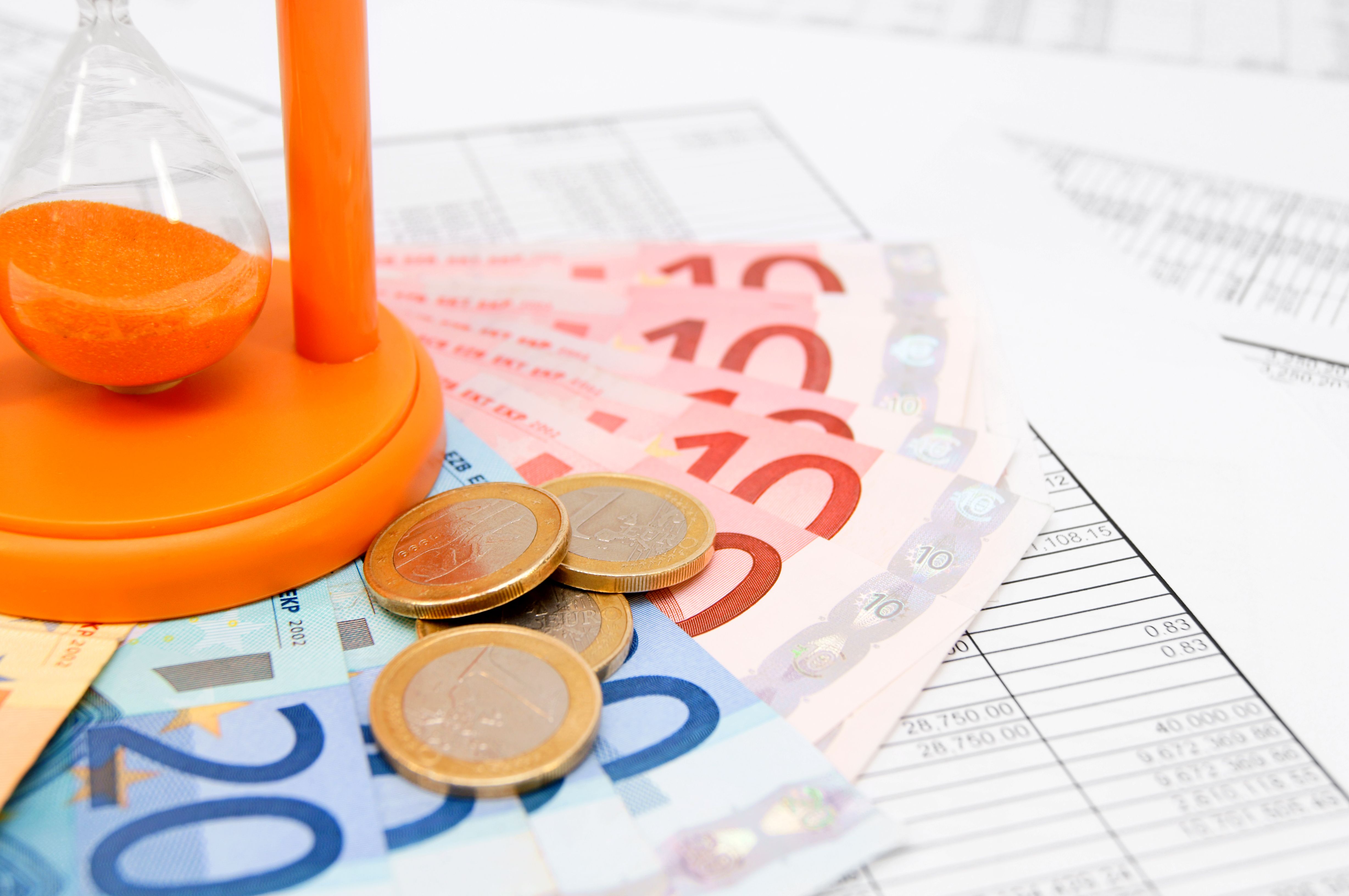 This photograph depicts a close-up view of a pile of colorful Euro banknotes, including 5, 10, and 20 denominations, laid on a white desk.