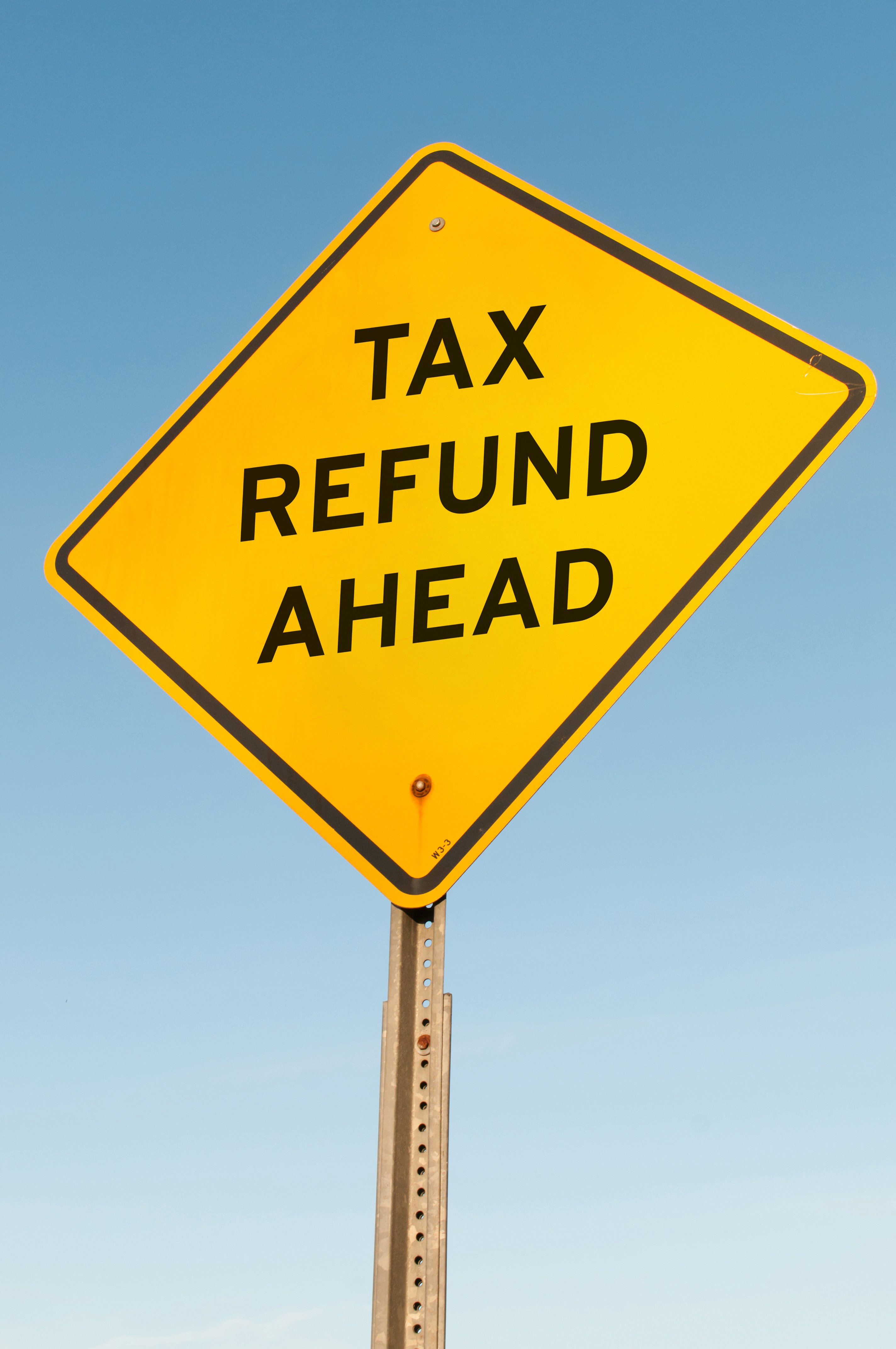 This is a photograph of a bright yellow diamond-shaped sign with a black border, mounted on a metal pole. The sign reads "TAX REFUND AHEAD" in bold, black uppercase letters.