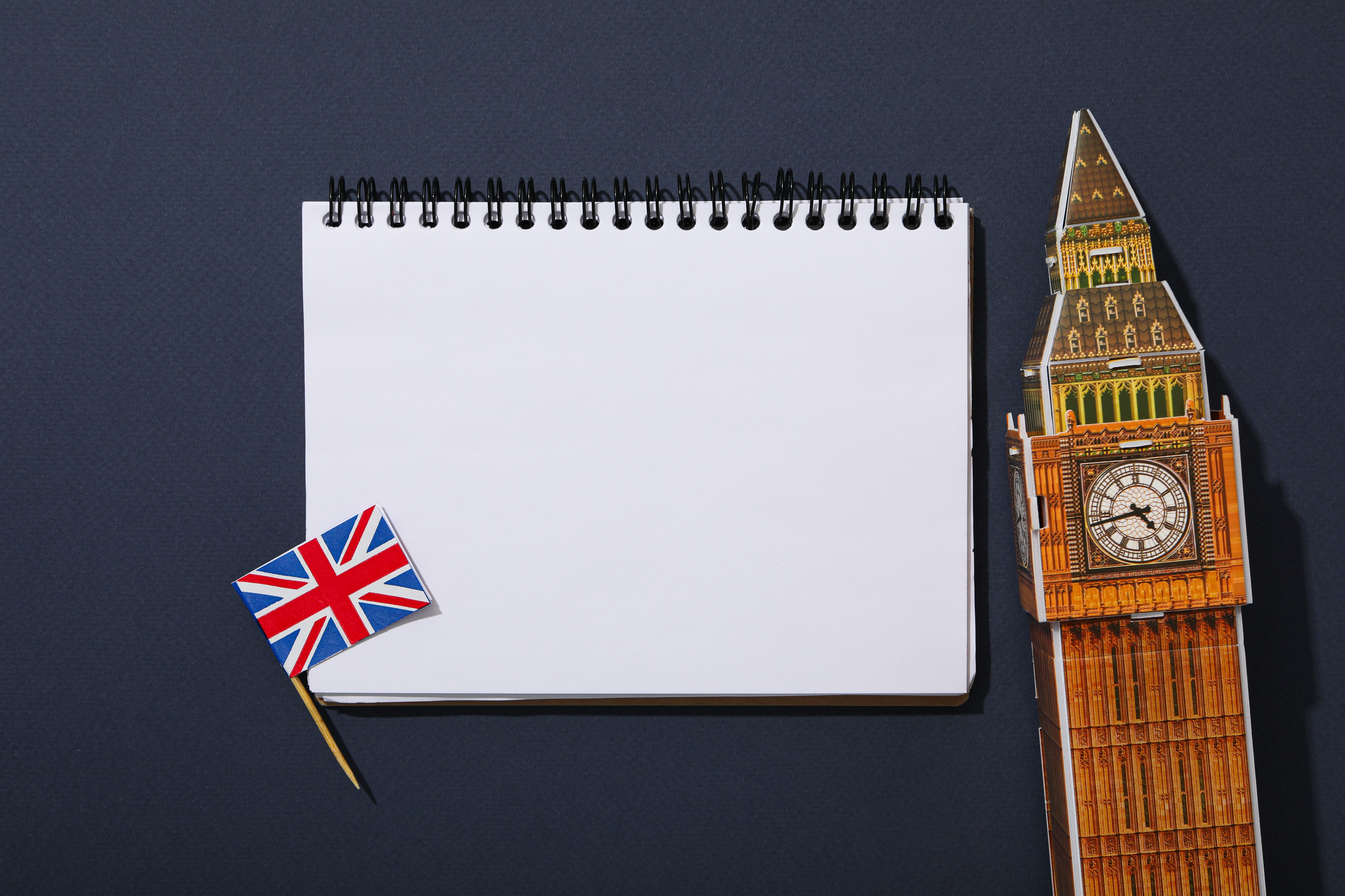 This photograph features a white spiral-bound notebook placed centrally against a dark blue background. The notebook is adorned with a small Union Jack flag pin on the bottom left corner. To the right of the notebook, a detailed, miniature model of Big Be