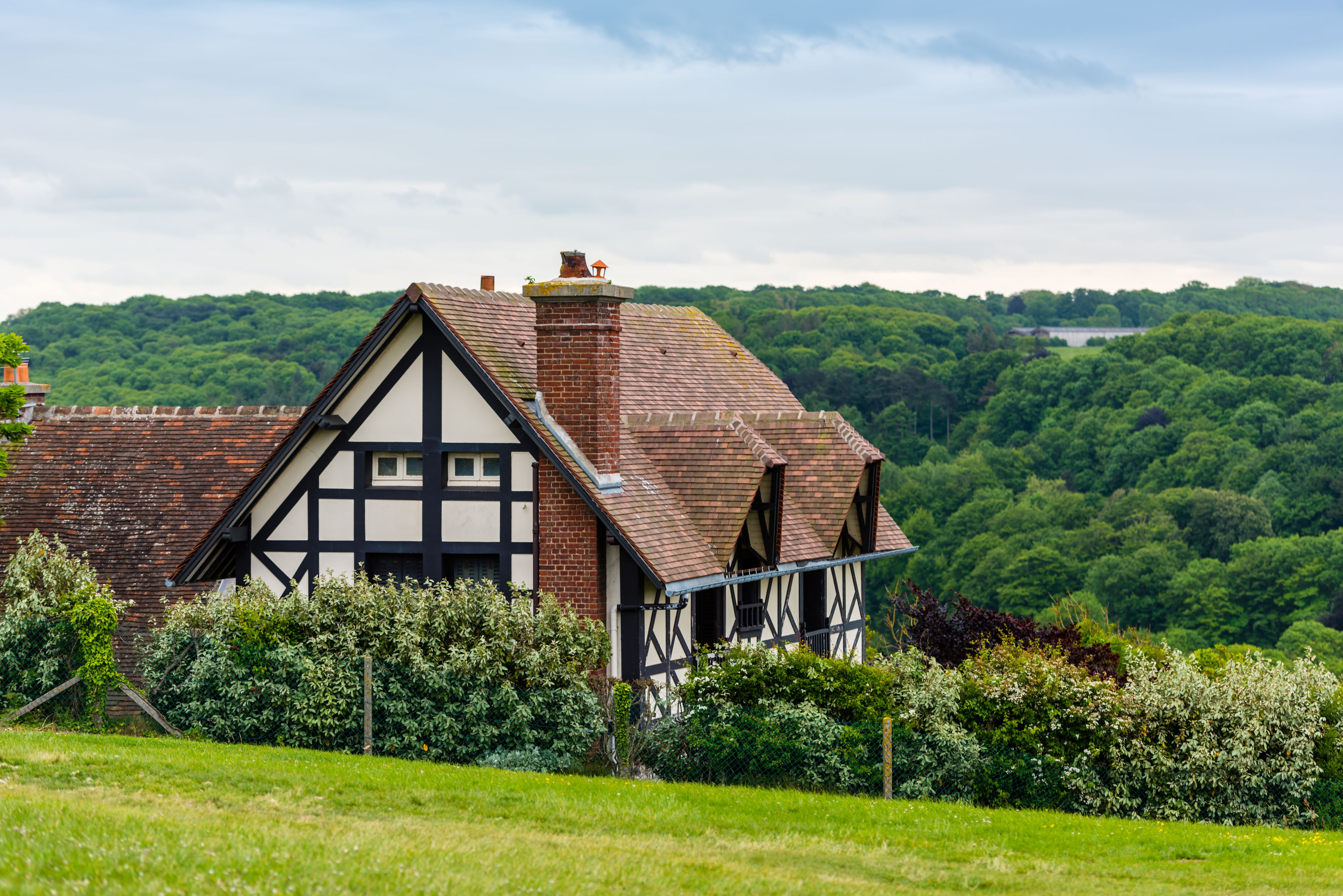 A photograph captures a charming Tudor-style house with dark wood framing and white plaster walls, set against a lush, green forest backdrop.