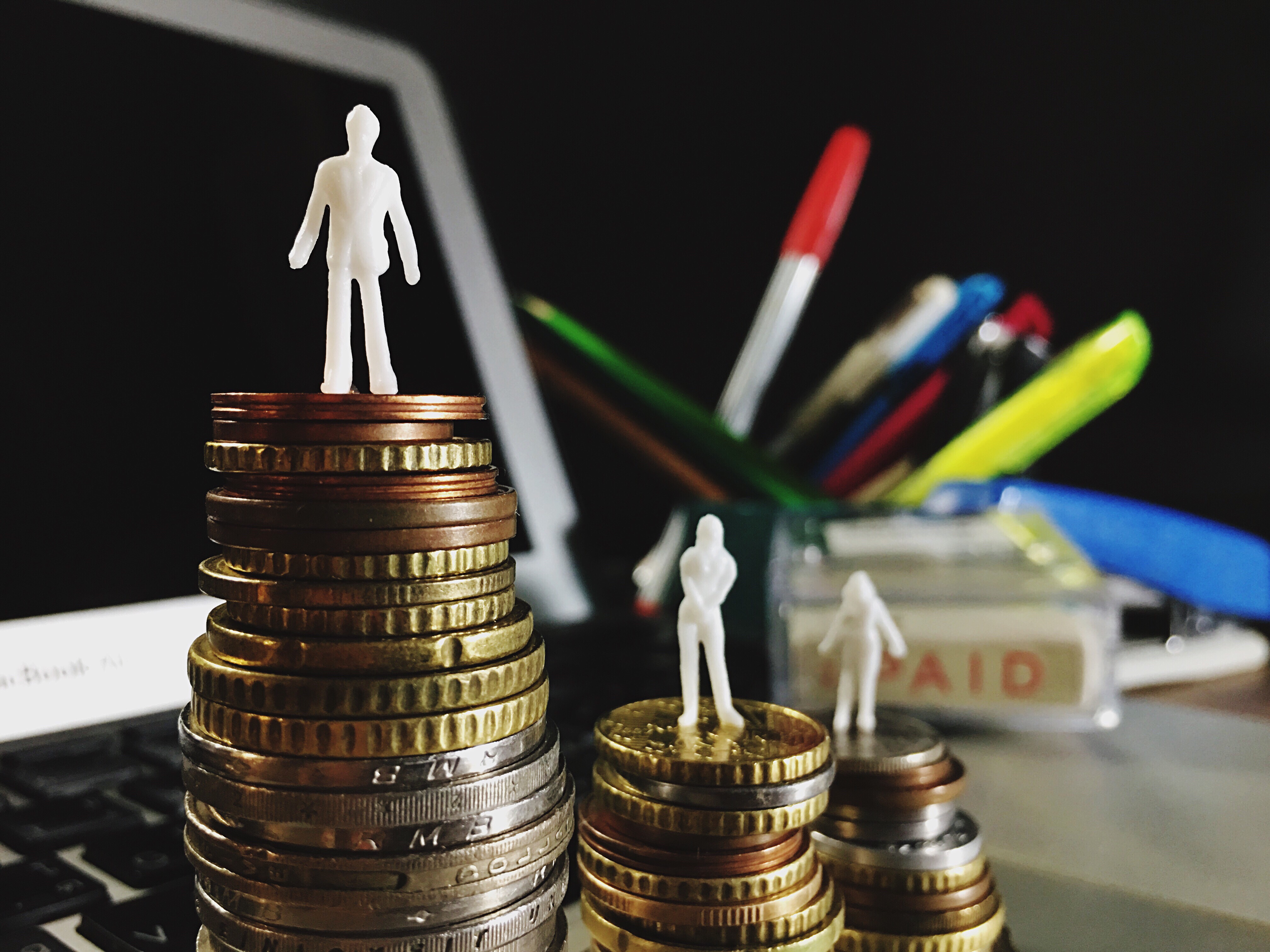 This is a photograph of a cluttered desk. In the foreground, three stacks of coins with small white human figurines on top, symbolizing wealth