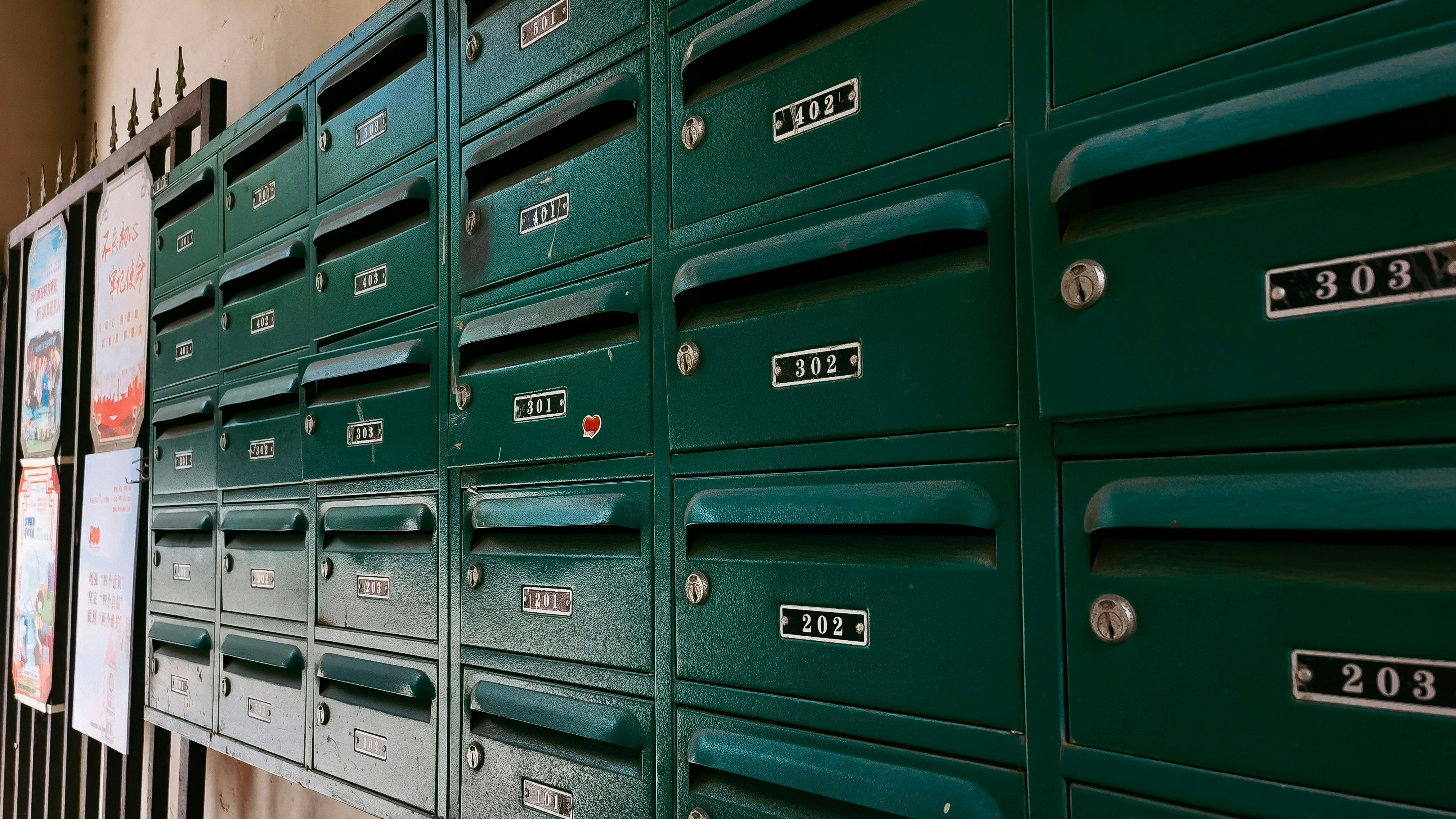 A photograph of a row of dark green, vintage metal mailboxes with white numbers and small red flags.