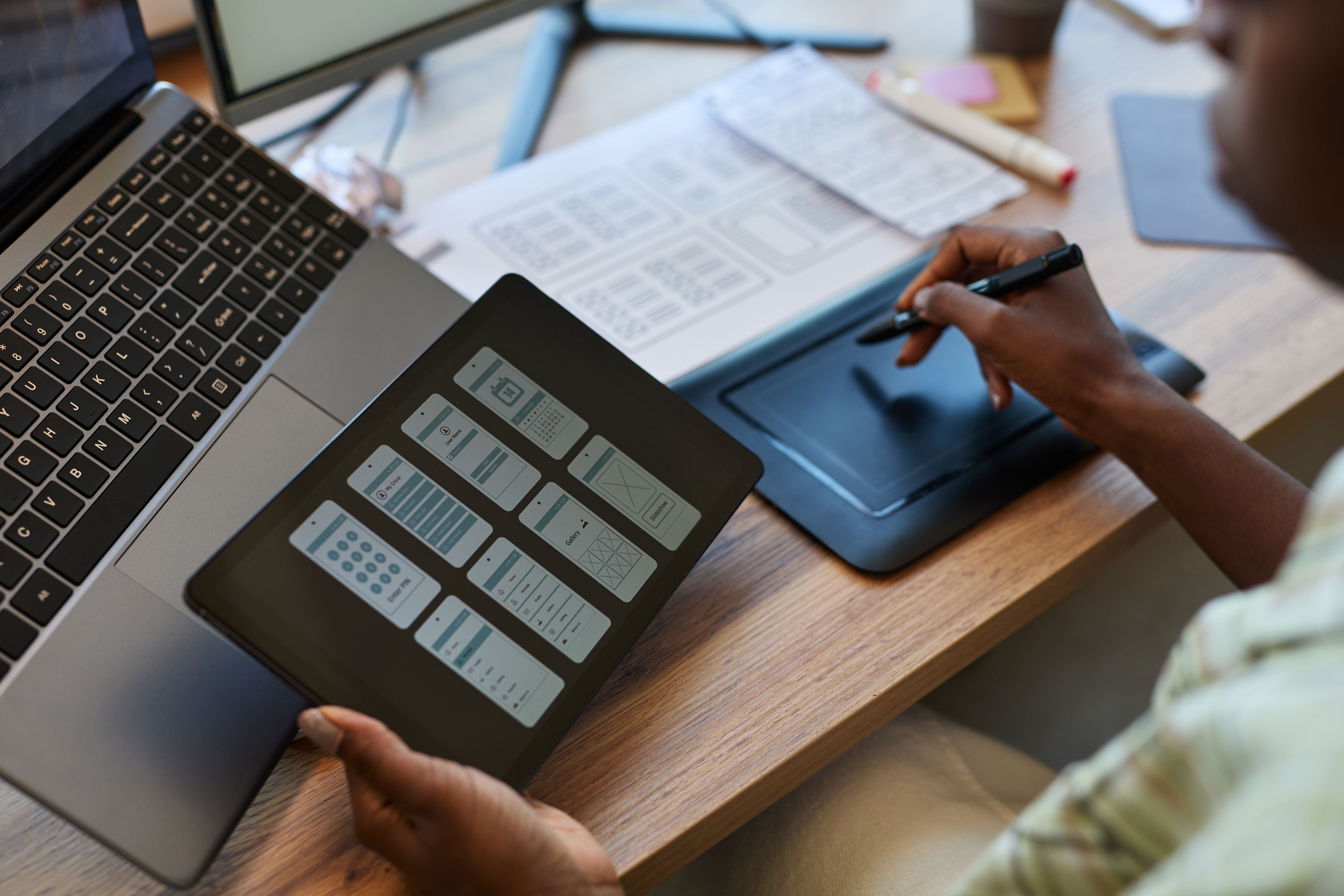 Photograph of a dark-skinned person, likely male, with a slim build and wearing a green plaid shirt, sitting at a wooden desk. 