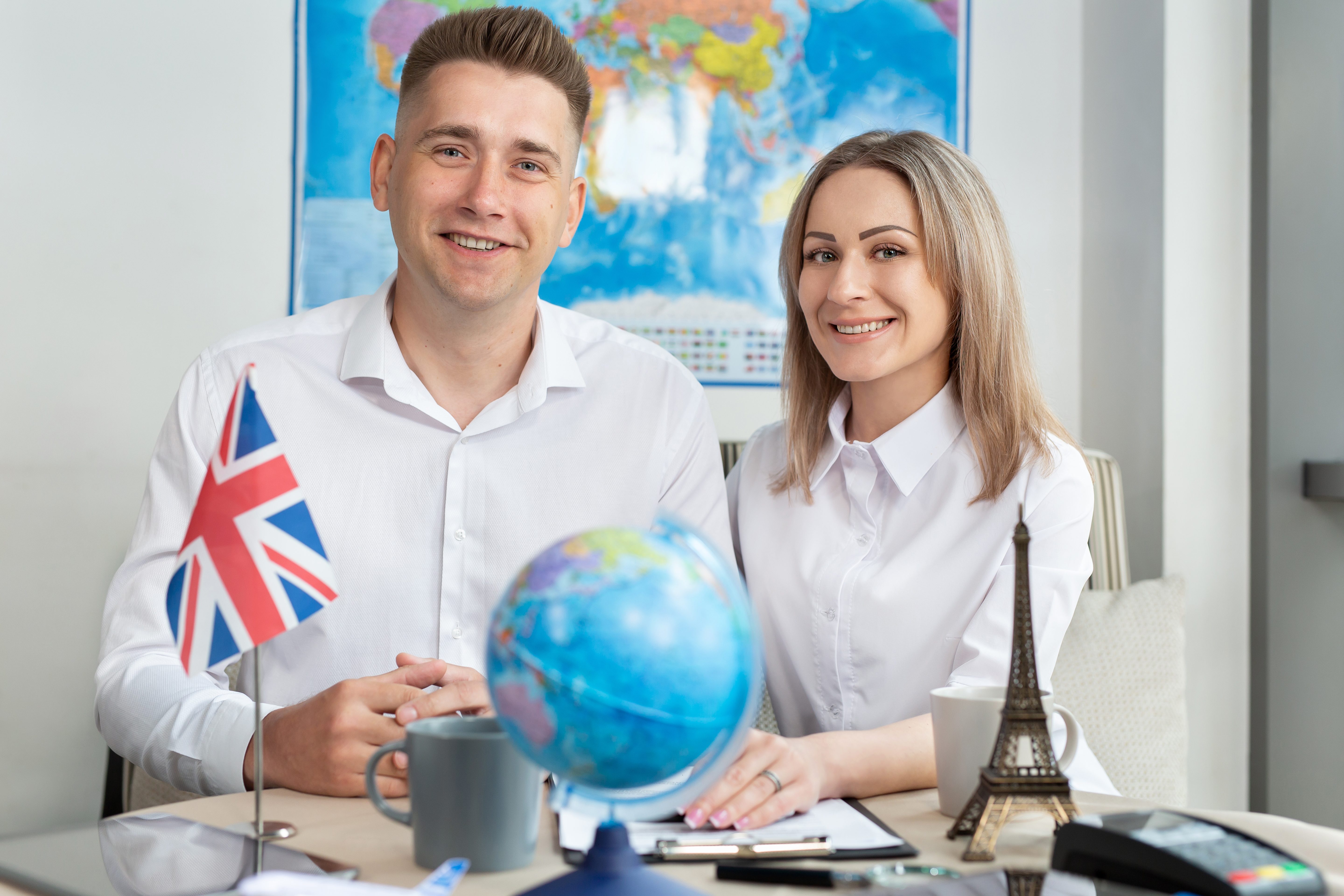Photograph of a Caucasian couple sitting at a desk. The man, with short brown hair, wears a white dress shirt and has the UK flag on his left shoulder. 